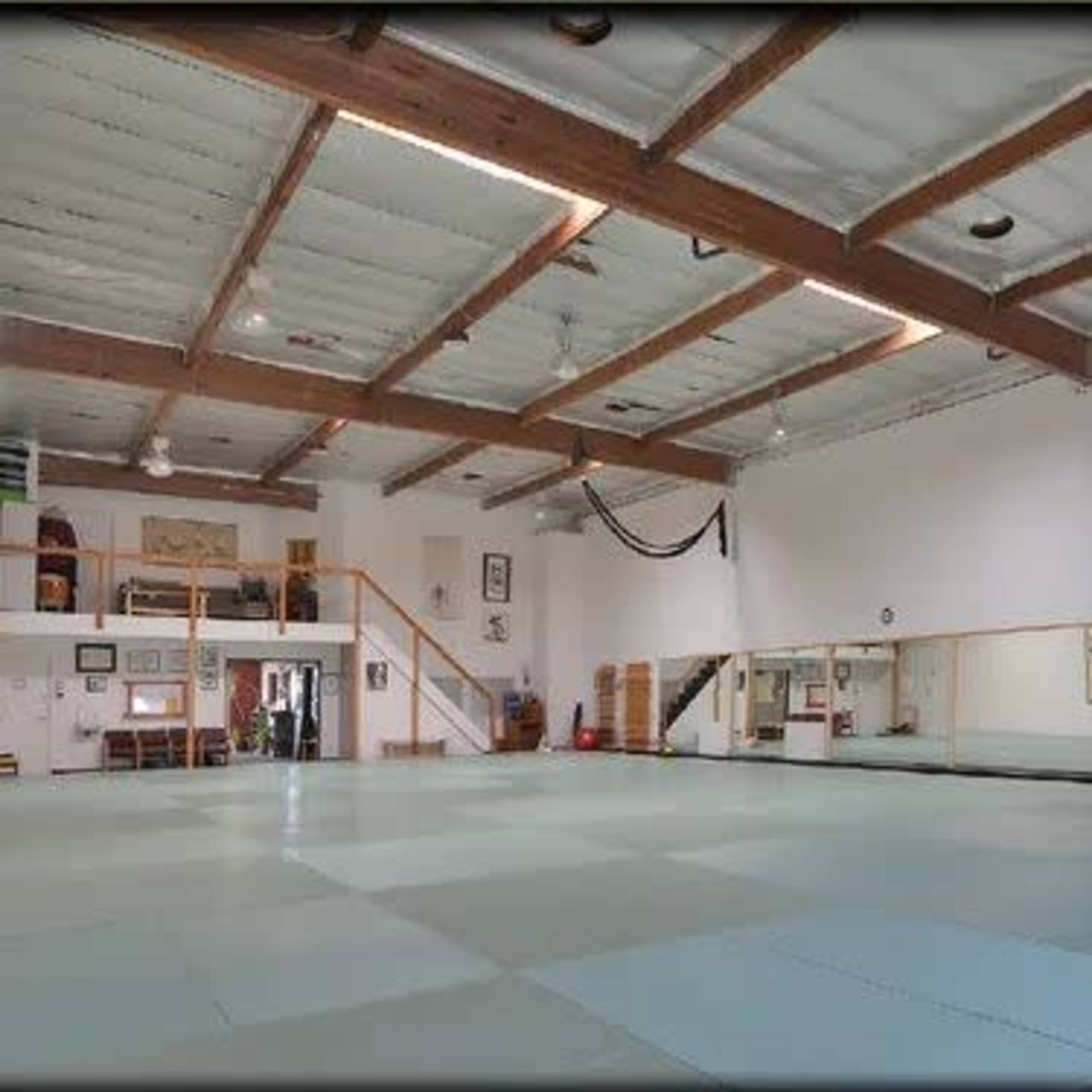 A spacious martial arts dojo with wooden beams, a mirrored wall, and soft practice mats on the floor.