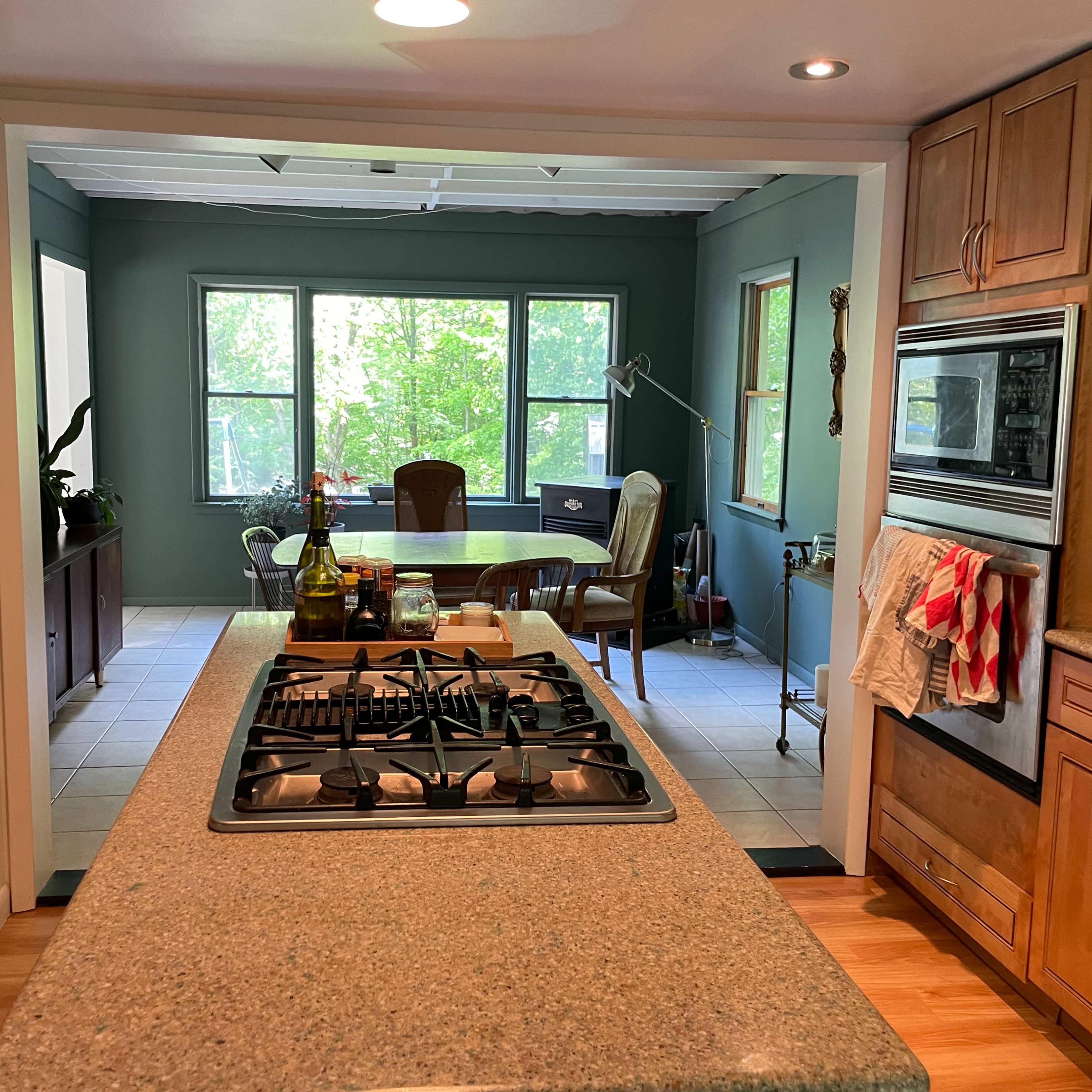 A kitchen with wooden cabinetry and a gas stove, leading to a dining area with large windows and a table set for meals.