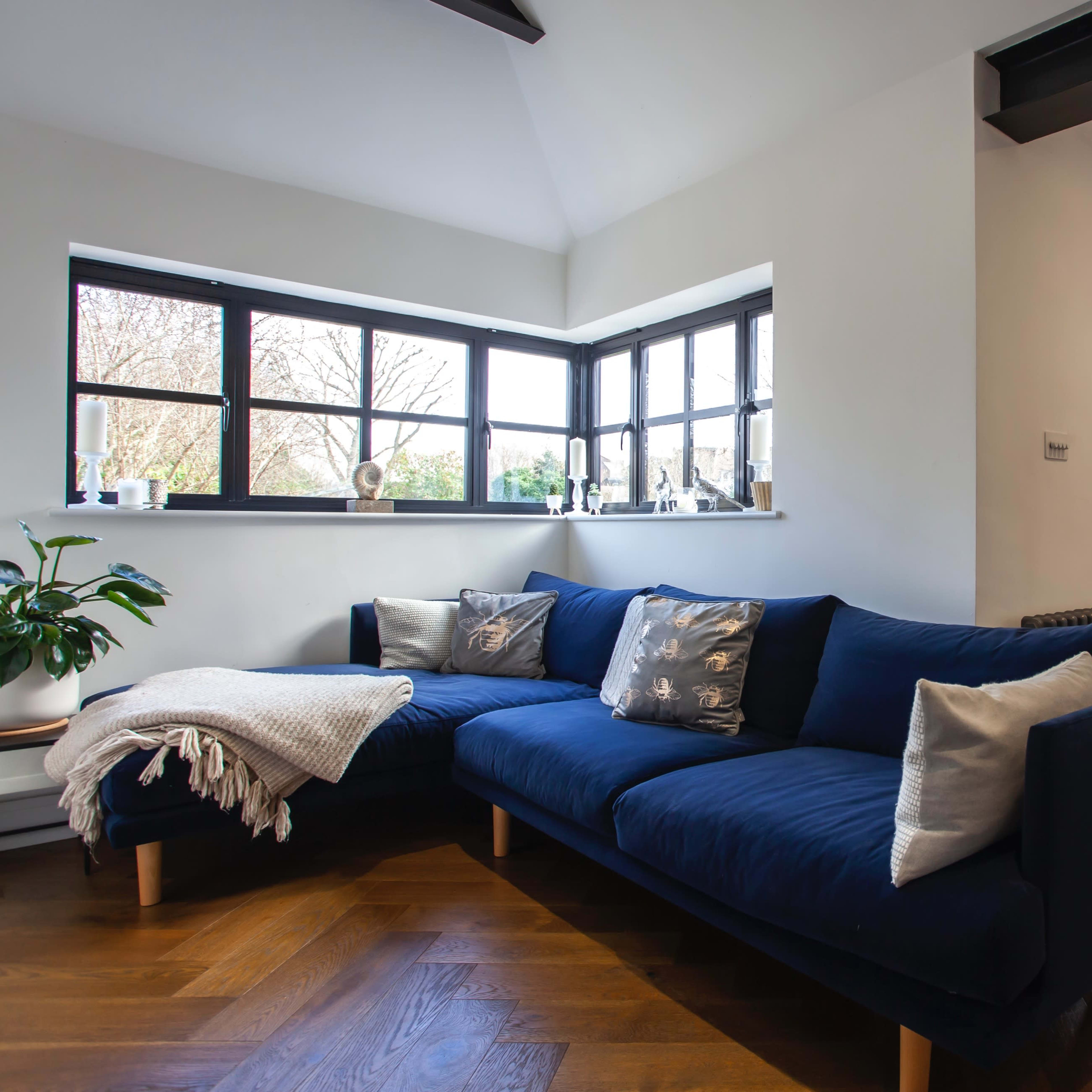 A modern living room features a navy blue sectional sofa, large windows allowing natural light, and a dining area with a wooden table.
