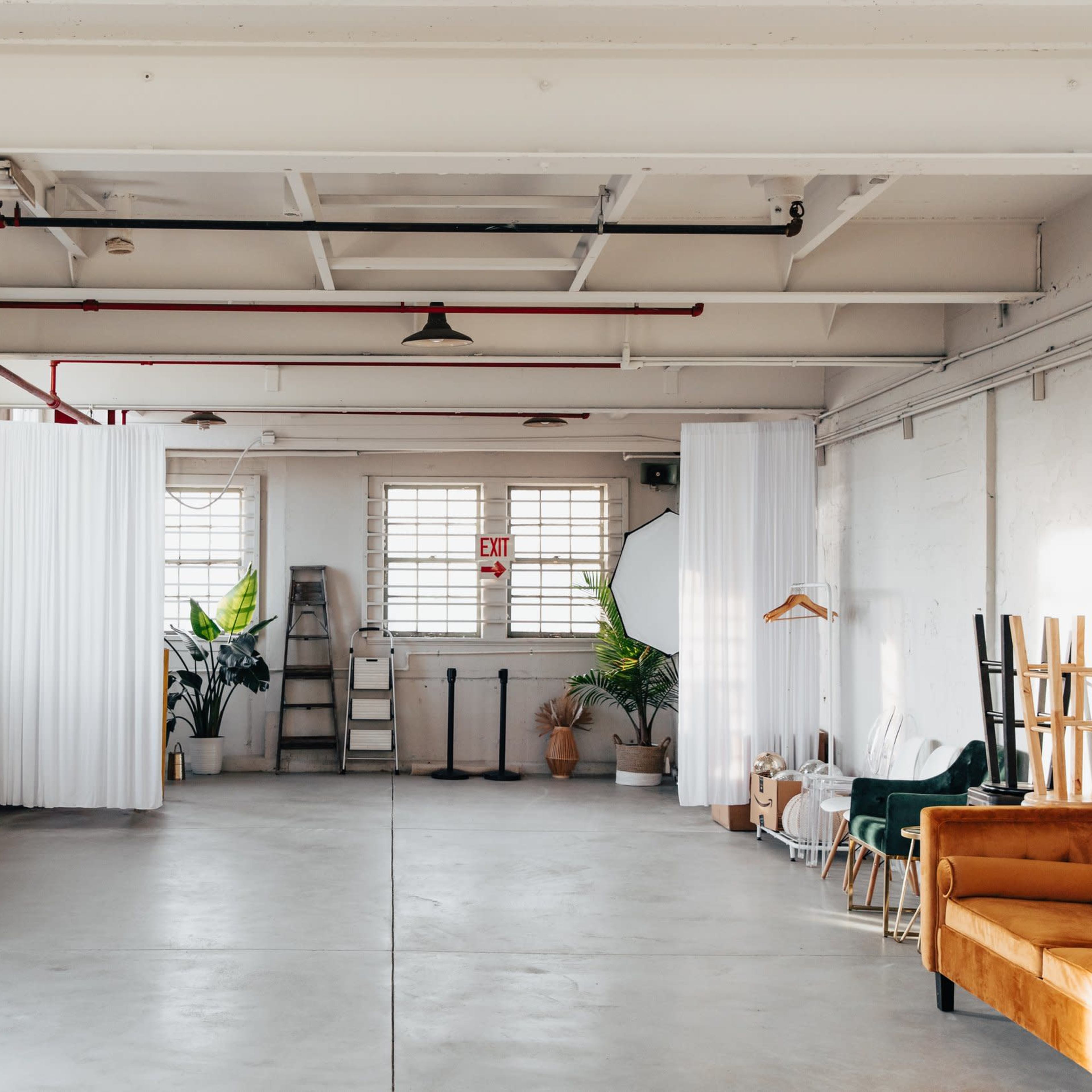A spacious, minimalistic room with white walls, concrete flooring, and large windows, featuring plants, light curtains, and seating arrangements along the sides.