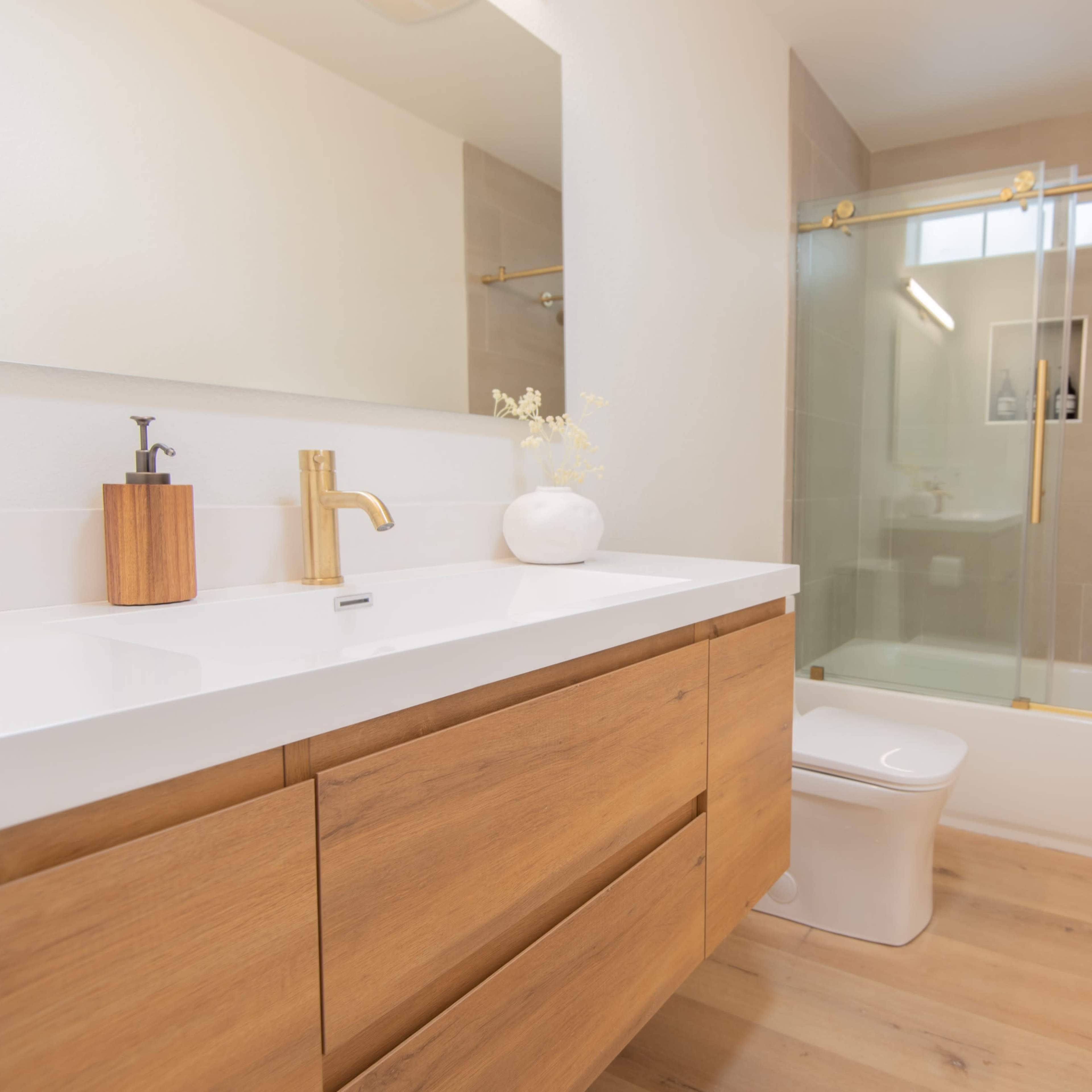 A modern bathroom featuring a wooden vanity with a white countertop, a toilet, a glass shower enclosure, and light-colored walls.