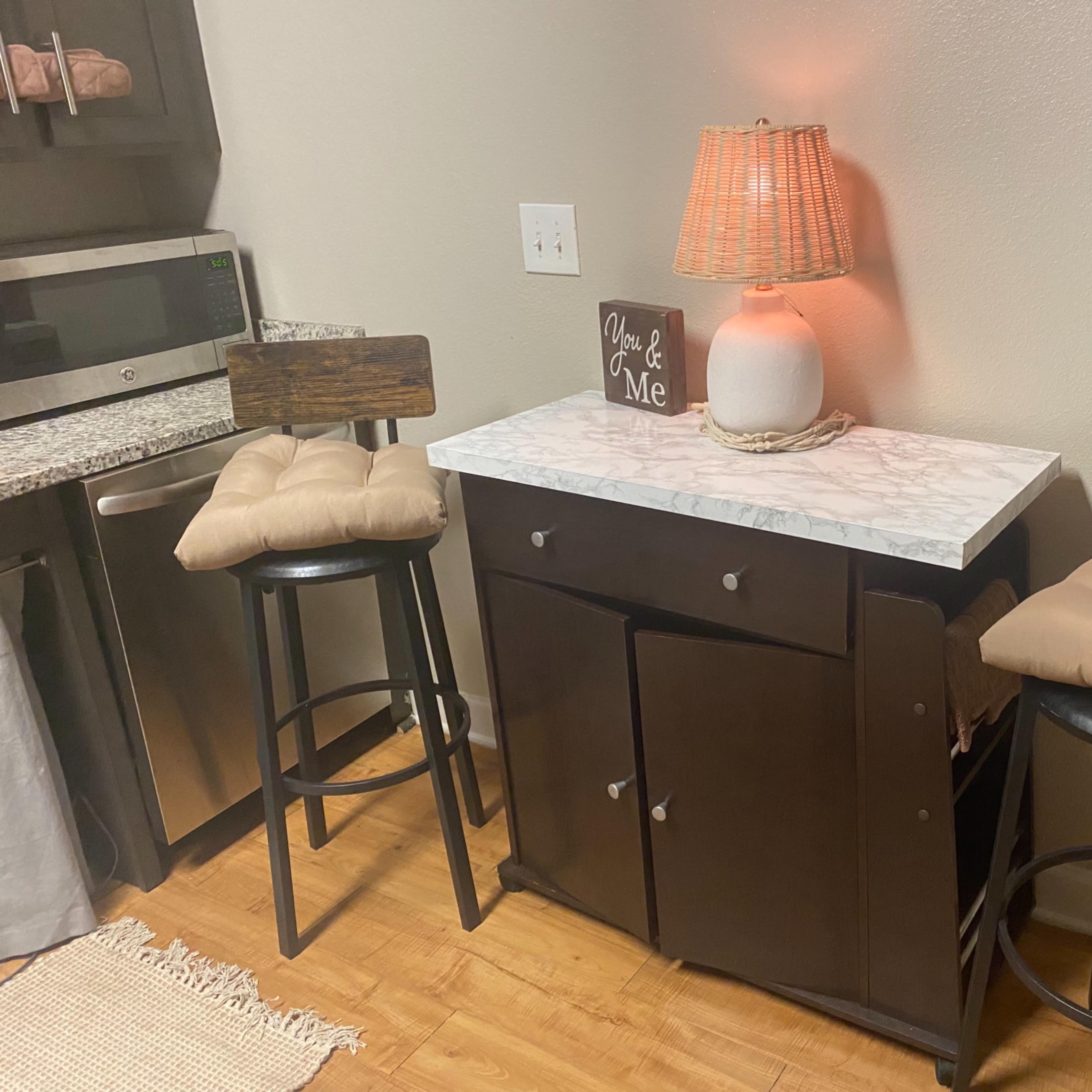 A small kitchen area featuring a side table with a marble top, two black stools with cushions, and a lamp with a woven shade.