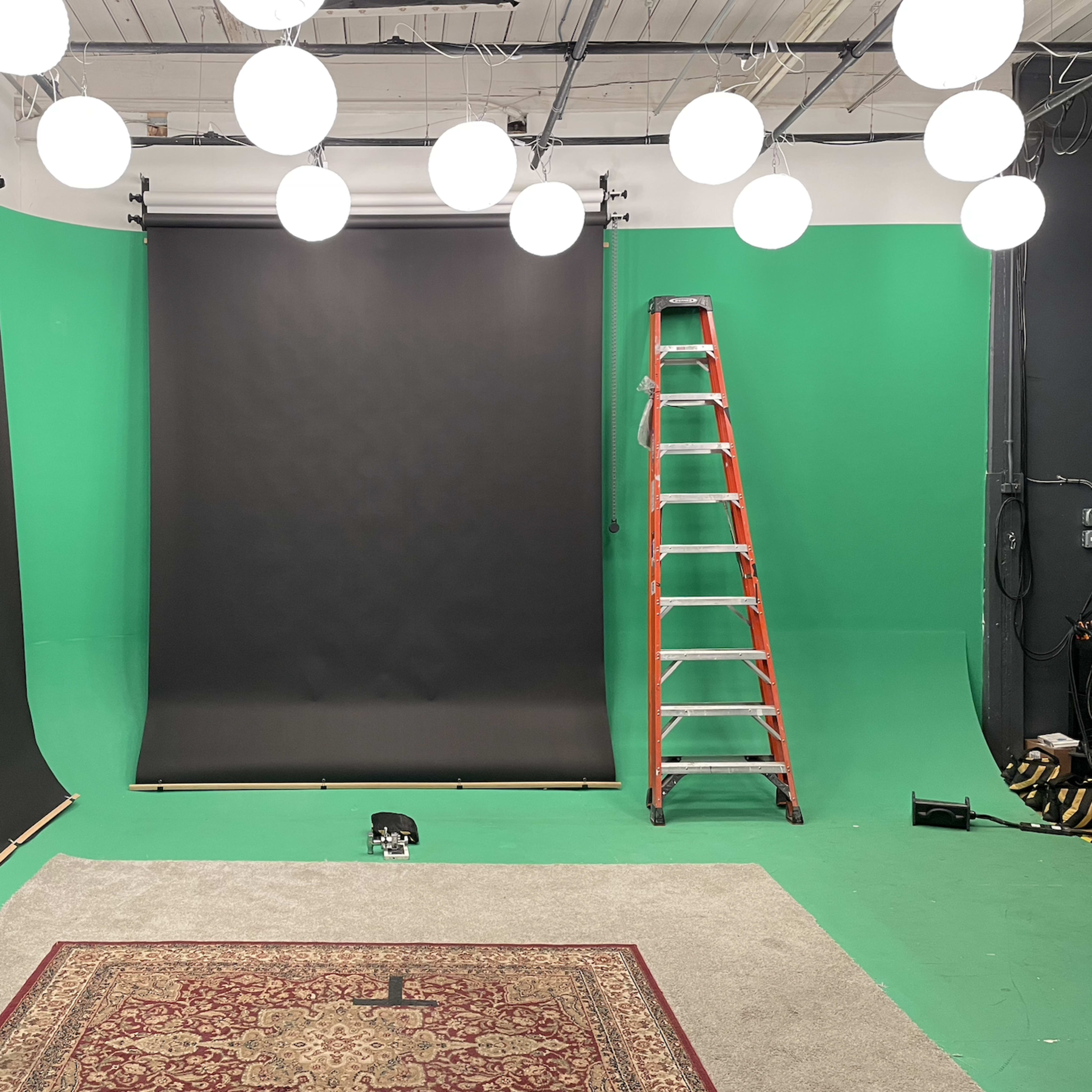 The scene features a green screen studio with two black backdrops, a ladder, and hanging lights above a carpeted area.