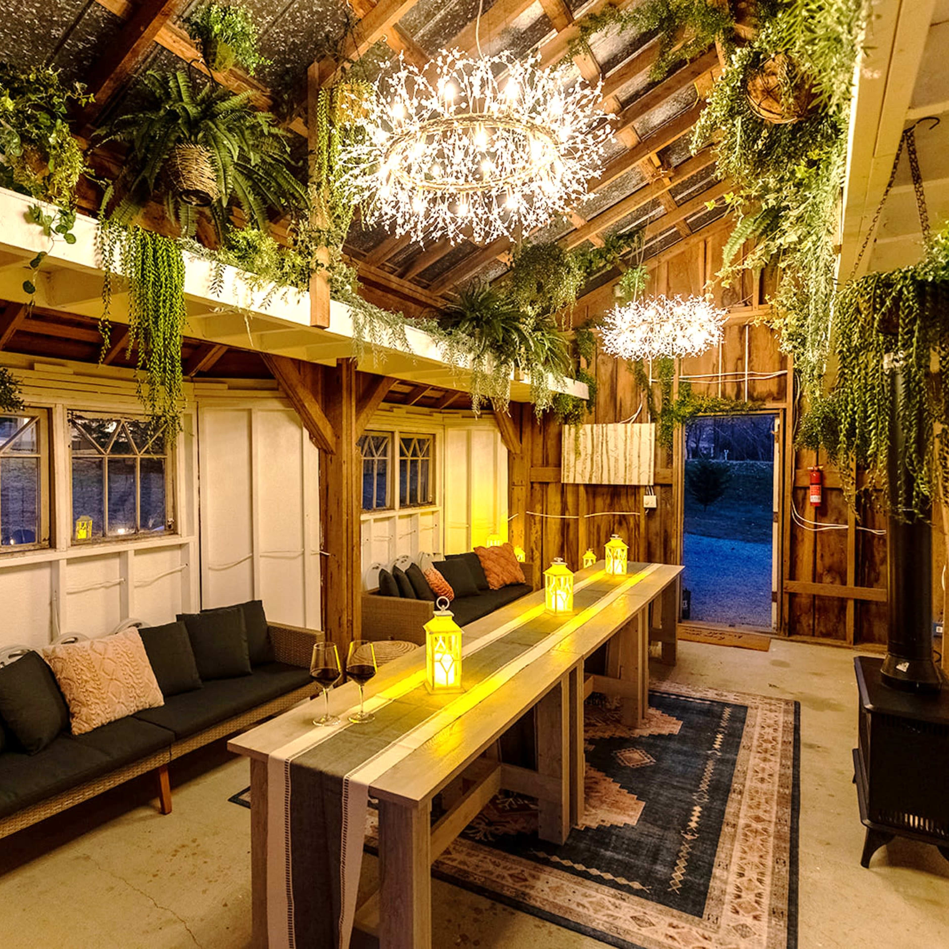 A rustic indoor space featuring a long wooden table, two stylish seating areas, and hanging lights amidst an array of plants.