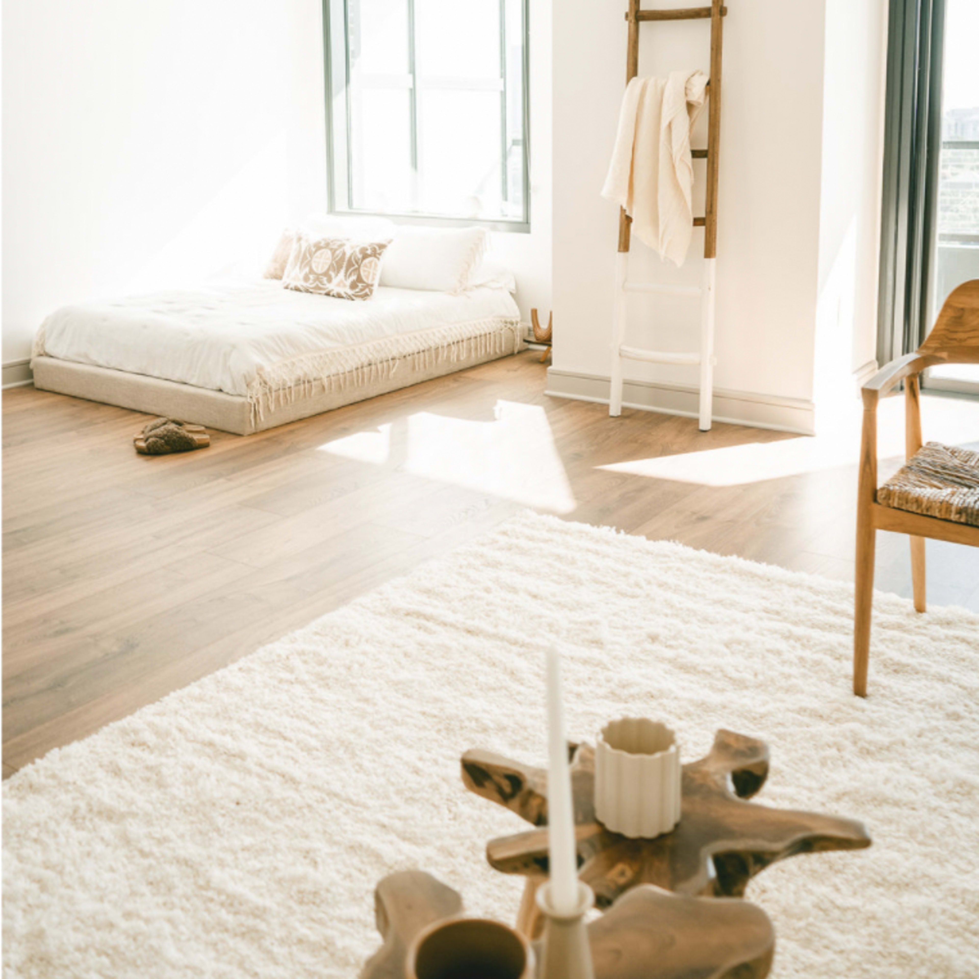 A minimalist room features a low mattress on the floor, a wooden ladder holding towels, and a cozy, textured rug.