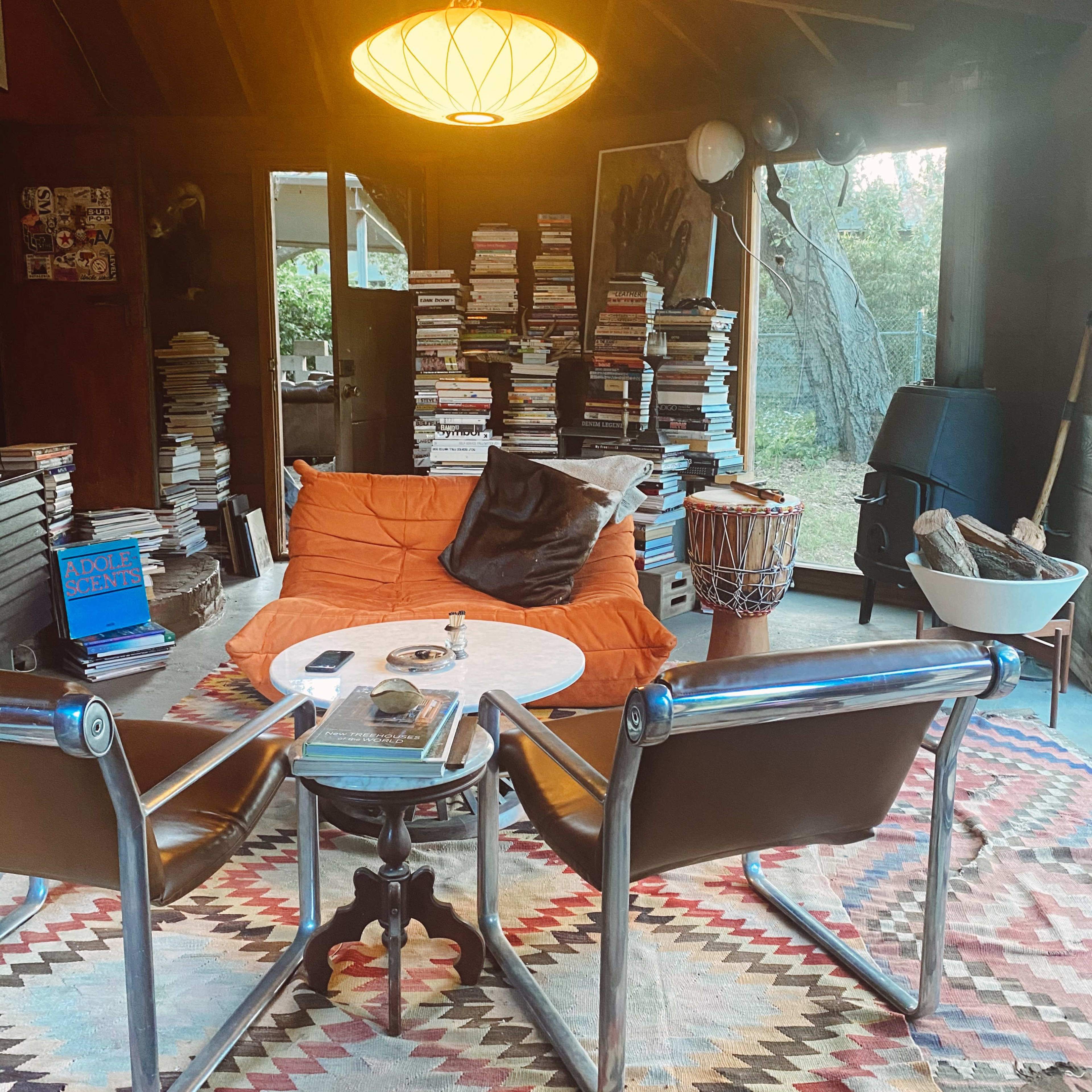 A cozy living room features a low table surrounded by chairs and a sofa, with stacks of books in the background and a large window letting in natural light.
