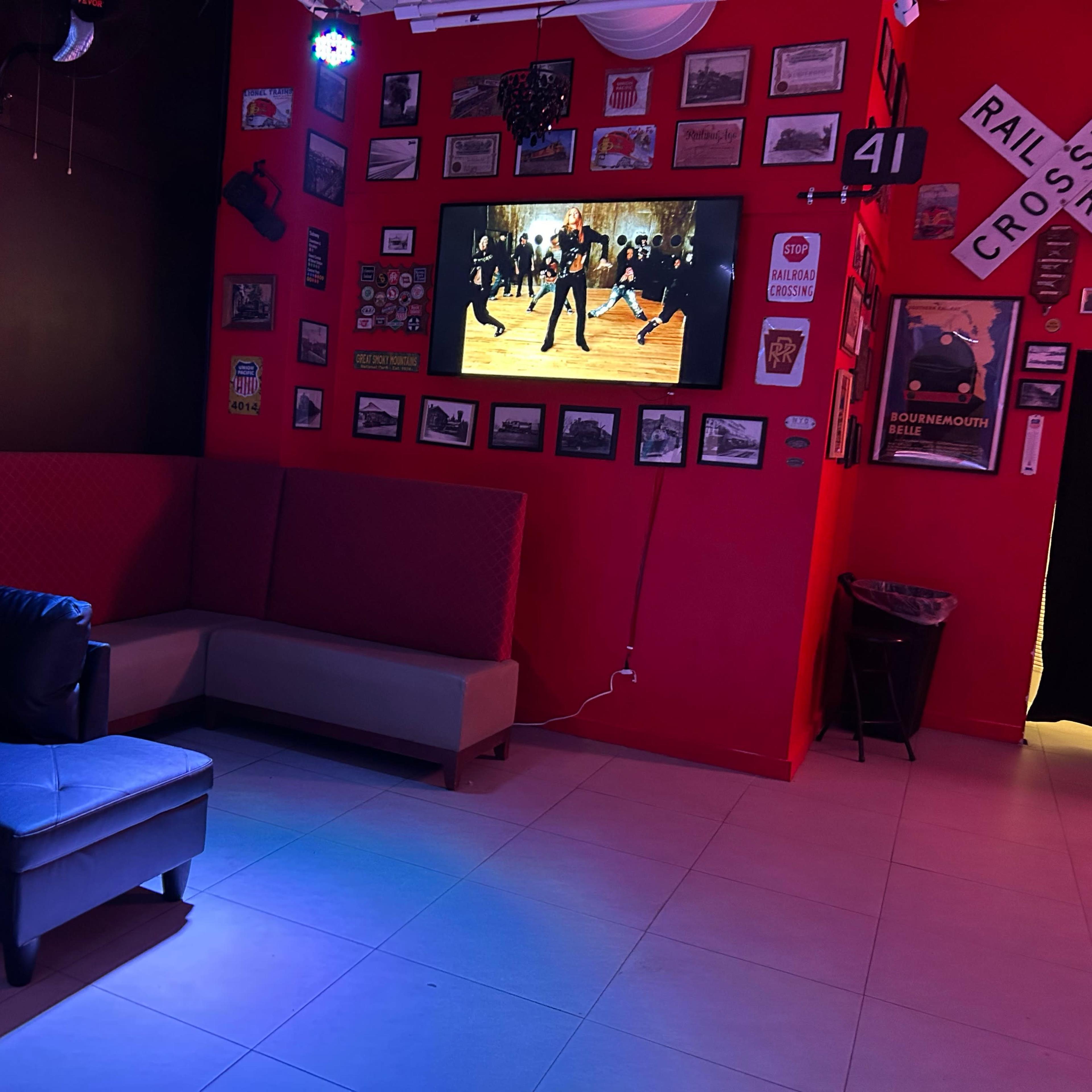 A lounge area with a dark red wall adorned with framed photographs and posters, featuring a large screen displaying a dance performance, a sectional couch, and ambient lighting.