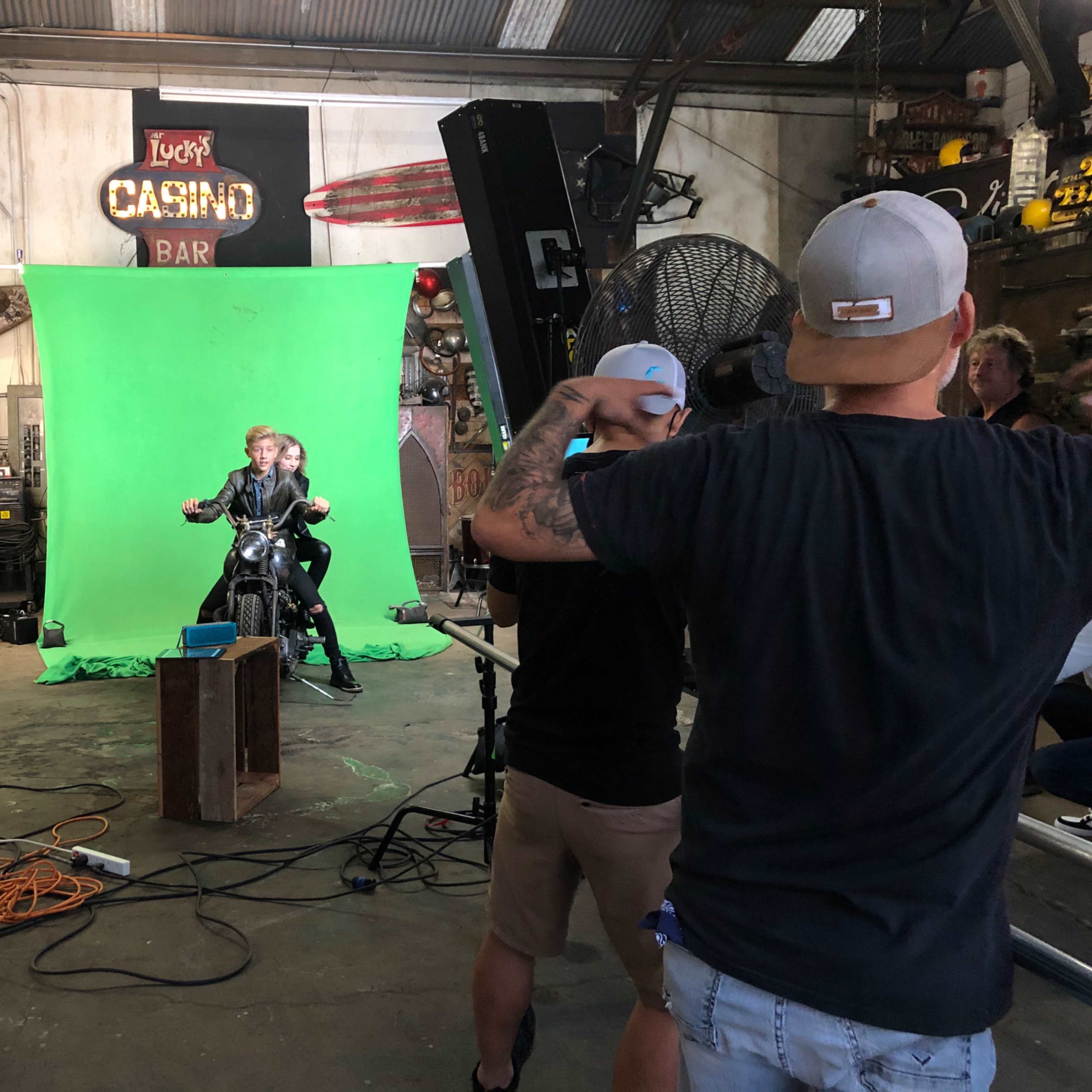 A film crew operates in a garage with a green screen while two individuals pose on a motorcycle.