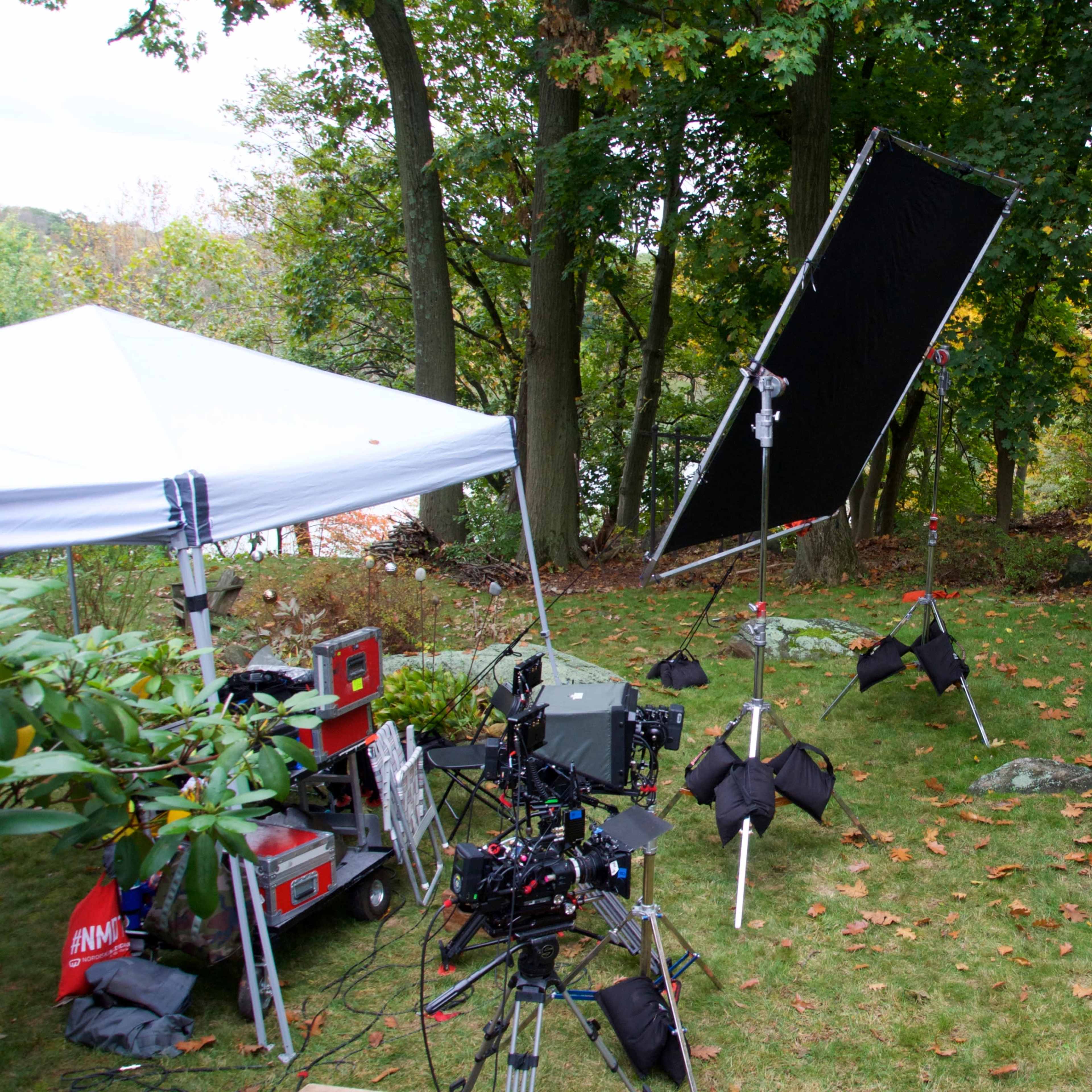 The scene features a film production setup in a wooded backyard, complete with a tent, lighting equipment, and an empty wooden bench.