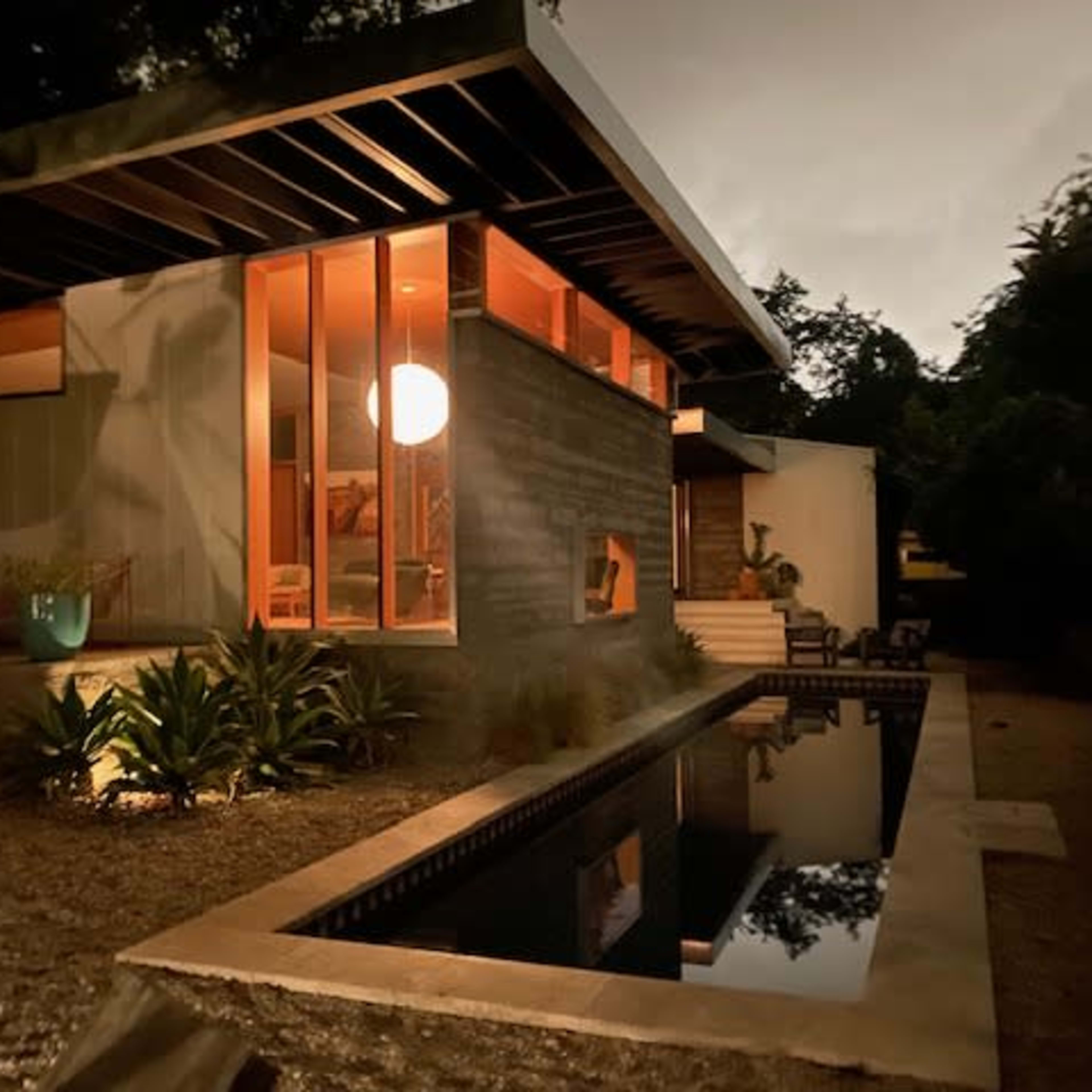 A modern house with large windows and a swimming pool reflecting the structure, surrounded by greenery in a dimly lit environment.