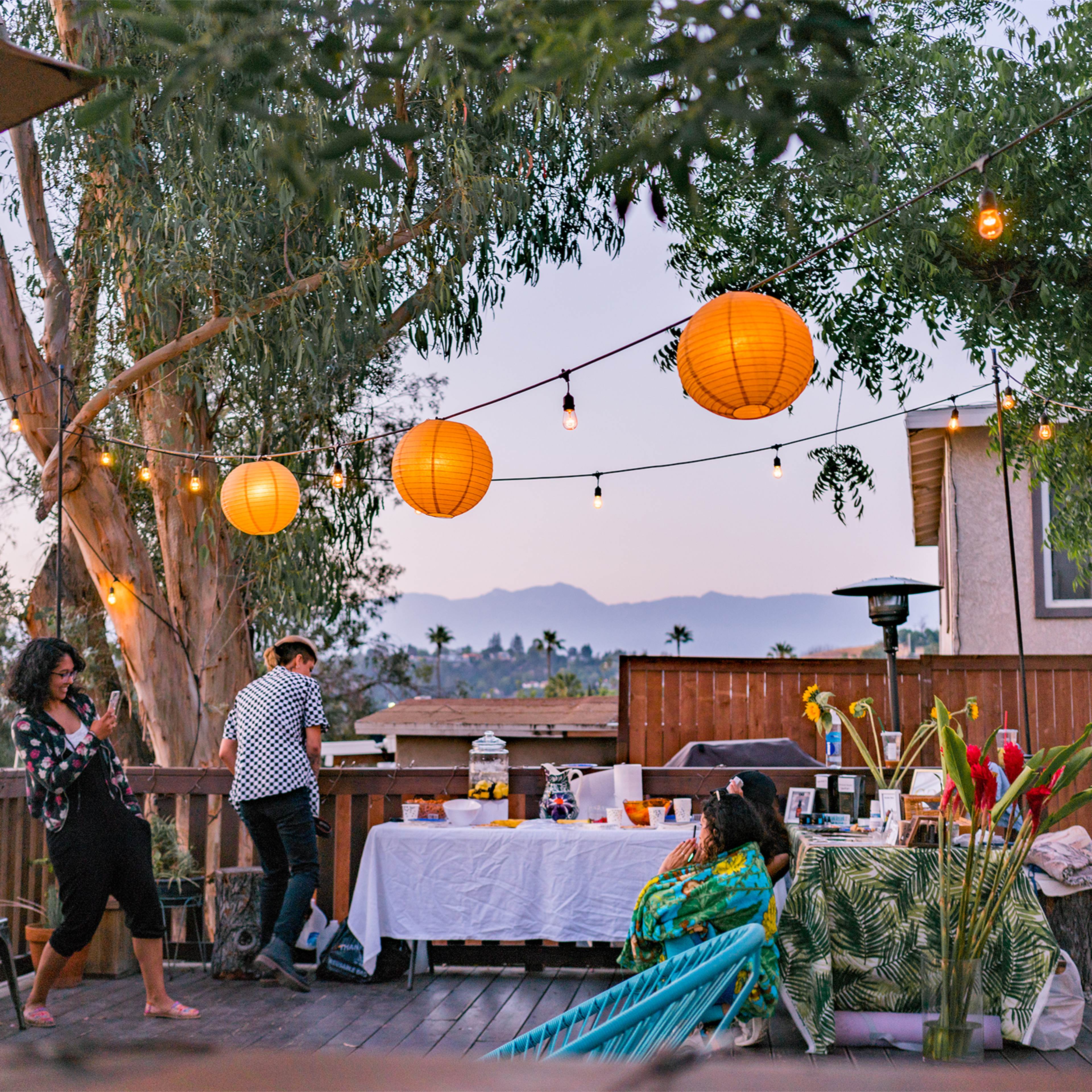 A group of people gather at an outdoor setting adorned with string lights and lanterns, sharing a meal as dusk approaches.