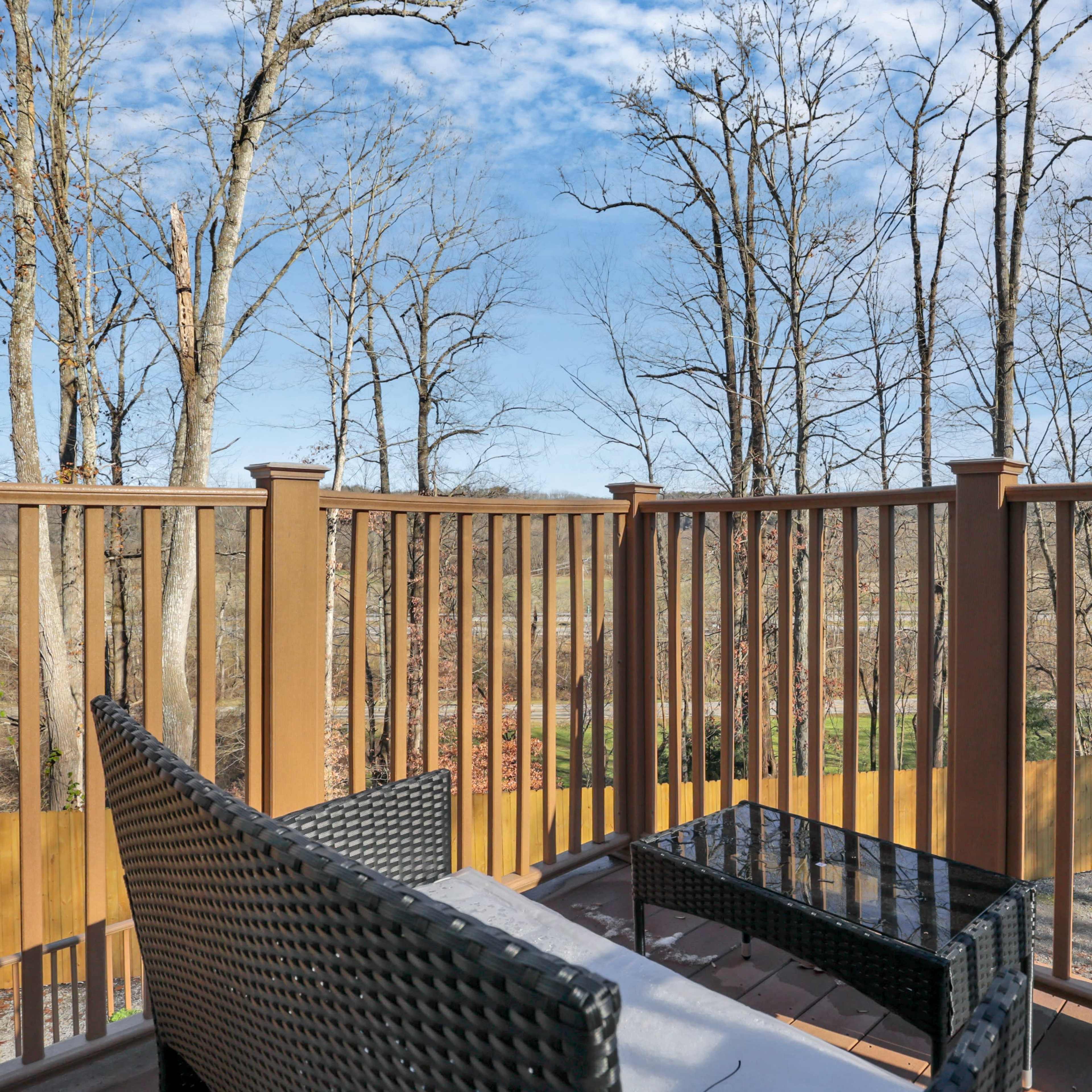 A wooden balcony with a woven seating area overlooks a wooded landscape with bare trees and a blue sky.