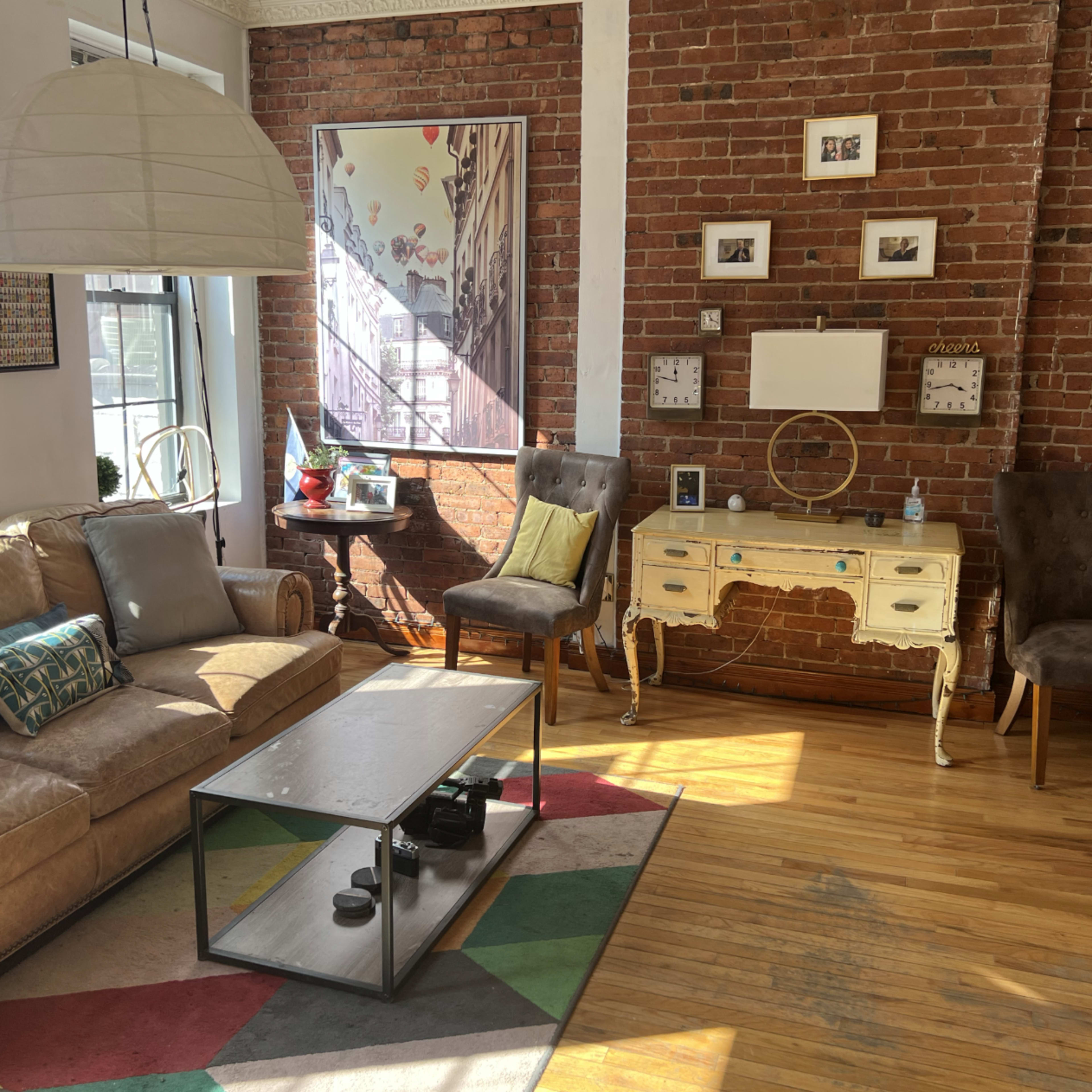A living room with a beige sectional couch, a coffee table, two upholstered chairs, and a console table against a brick wall.