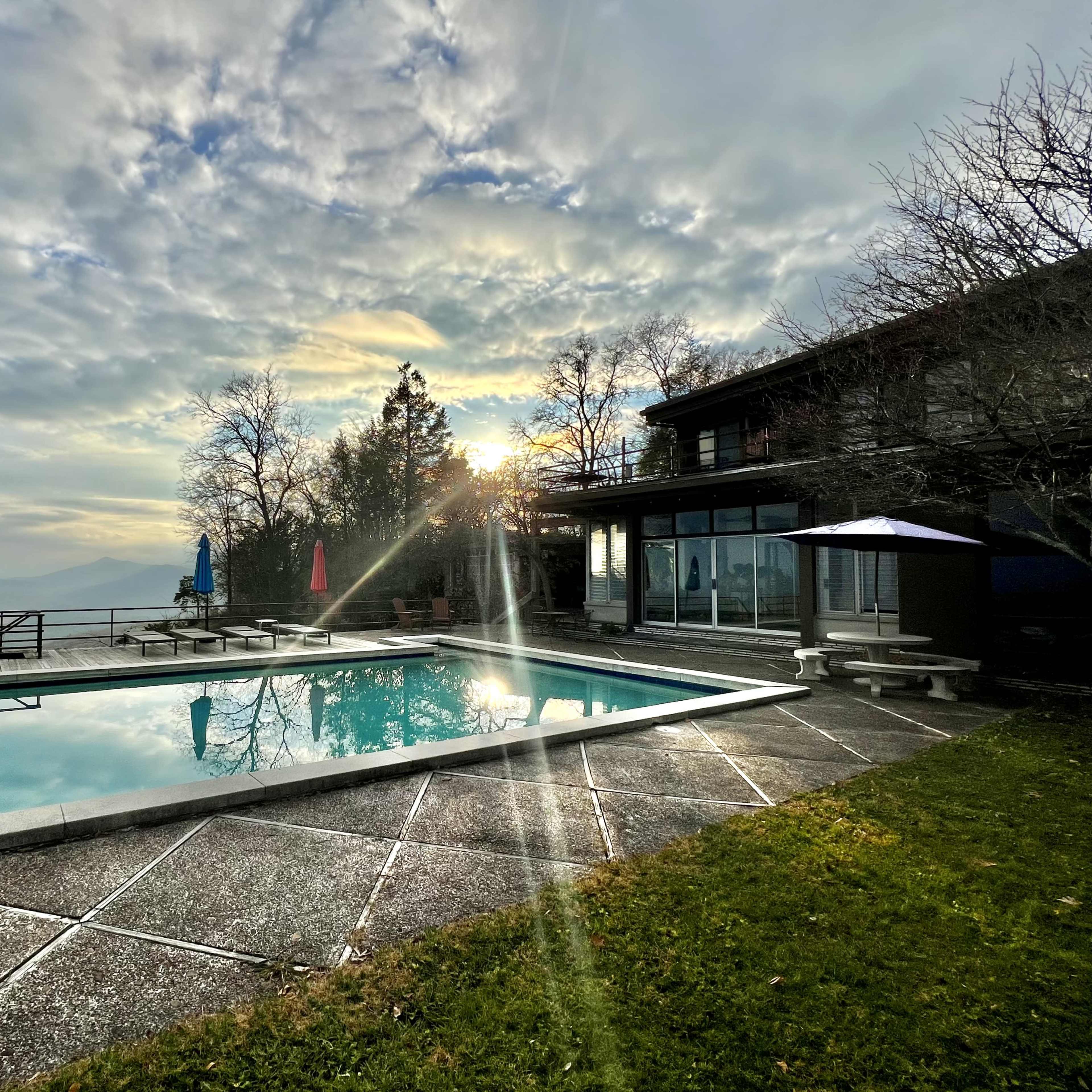 A rectangular swimming pool is situated beside a modern house with large windows, framed by trees and bathed in the light of a setting sun.