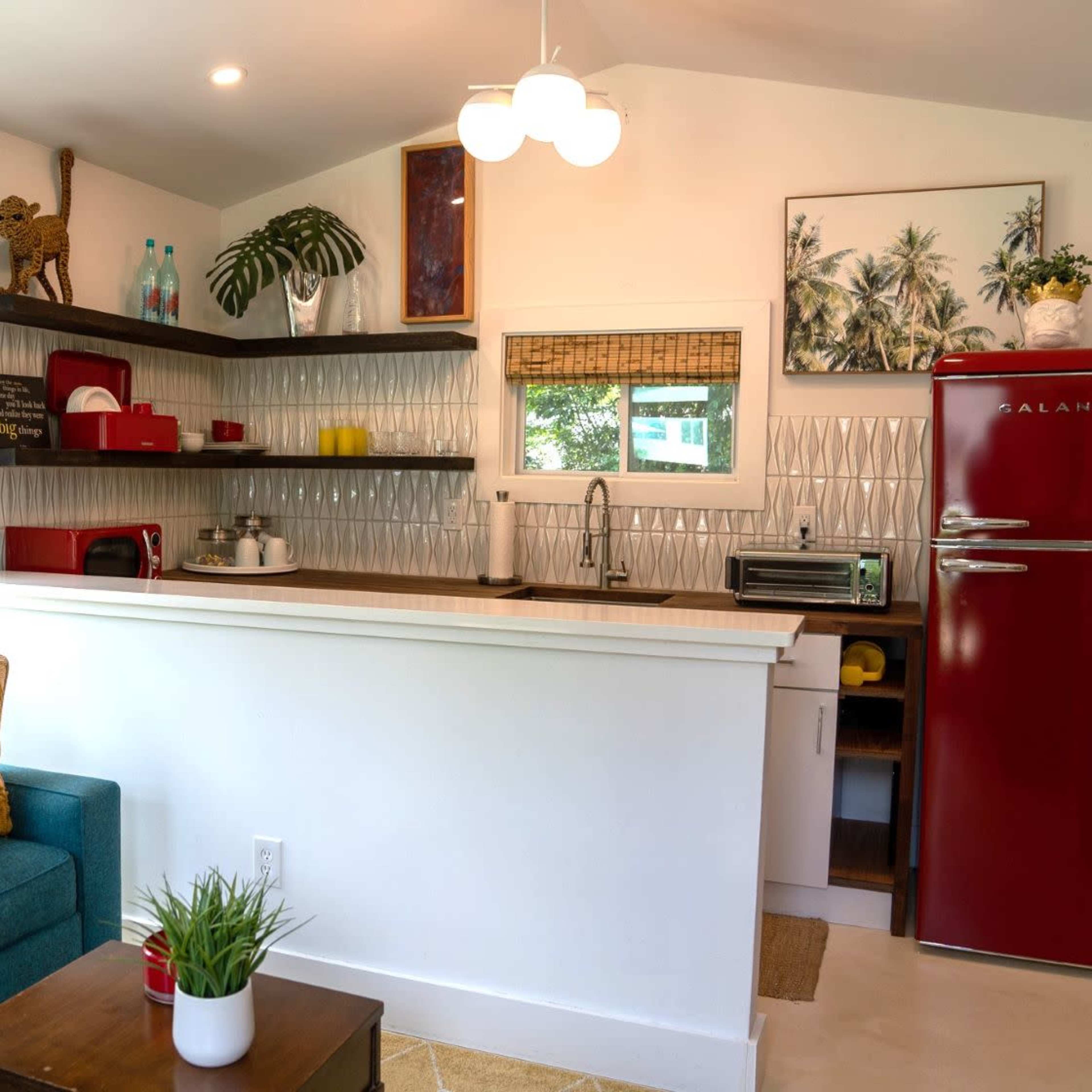 A compact kitchen and living area featuring a red refrigerator, a teal couch with a yellow pillow, and shelves adorned with various kitchen items and decor.