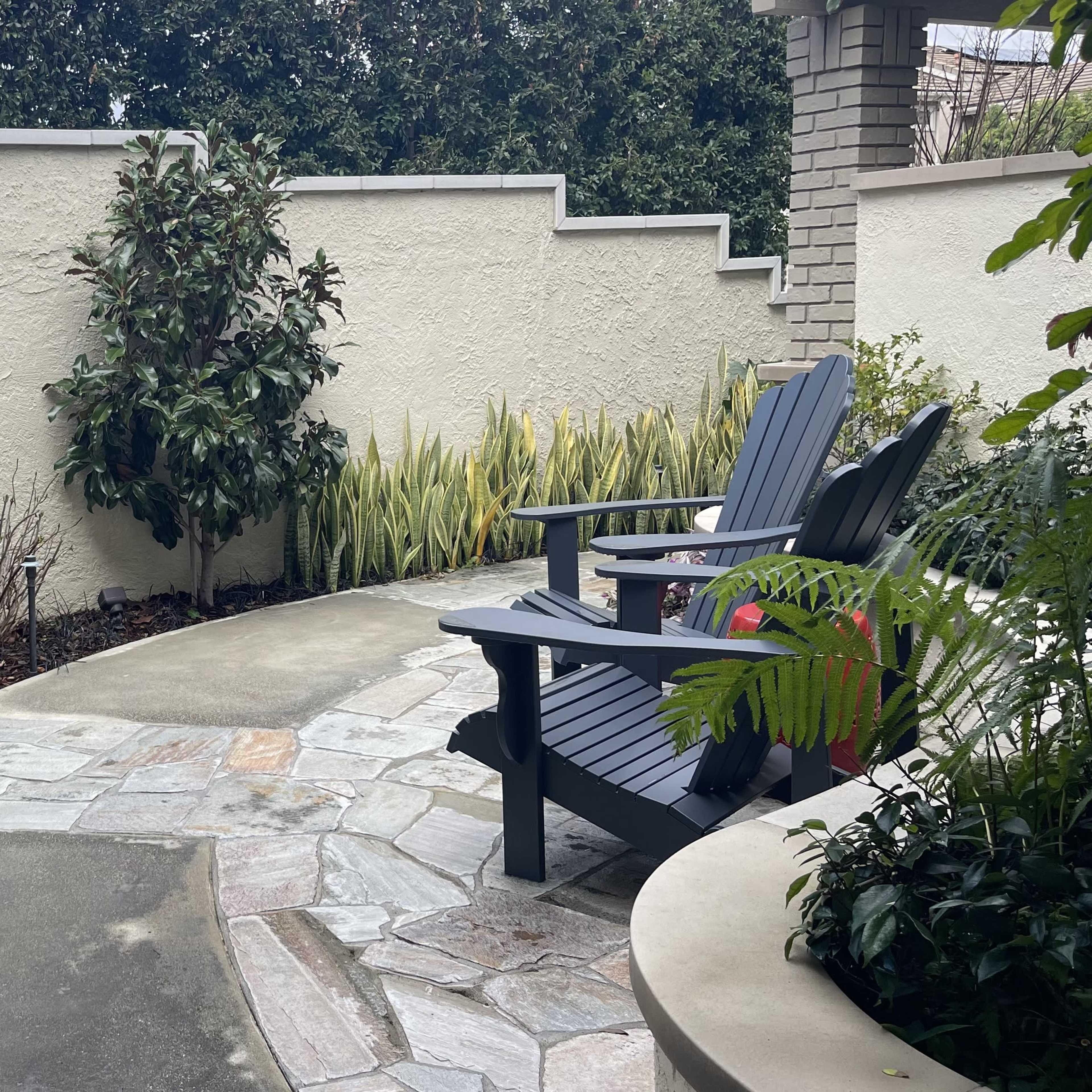 A landscaped garden area with two wooden chairs positioned beside a small stone birdbath, bordered by shrubs and plants.