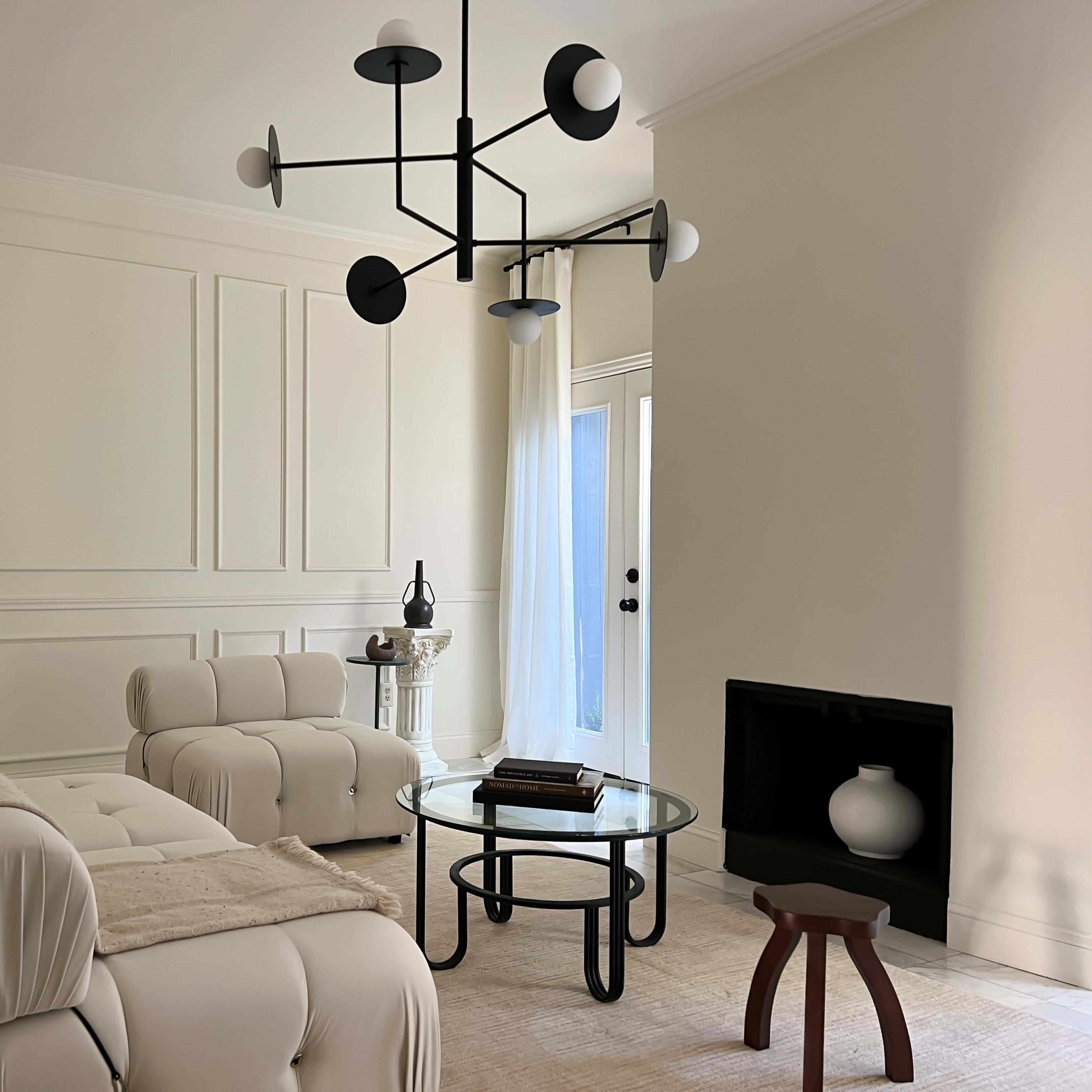 A minimalist living room features a white sectional sofa, a circular glass coffee table, and a modern chandelier, with a decorative sculpture and a black fireplace.
