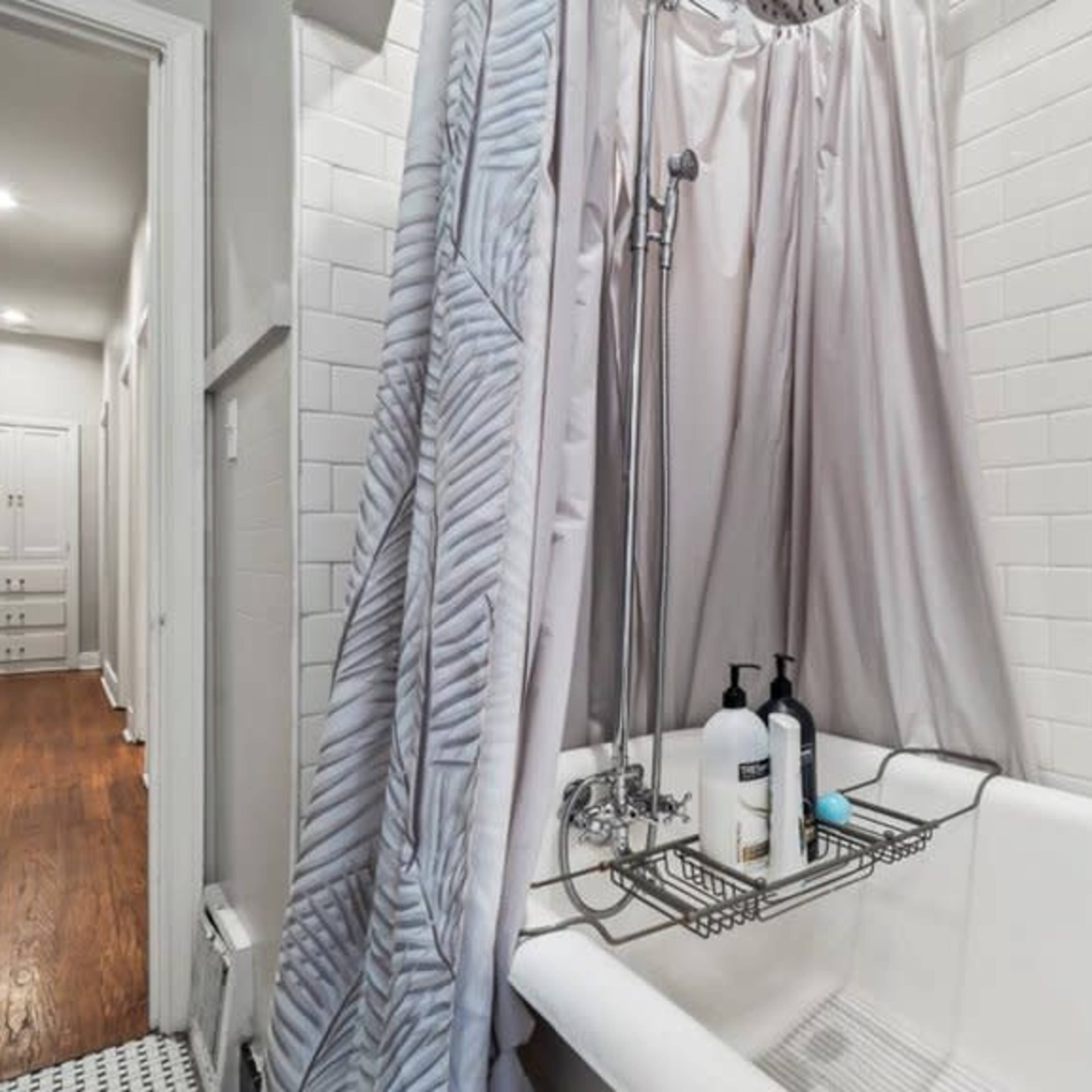 A bathroom with a bathtub and a shower curtain, featuring white tiled walls and a wooden floor visible in the adjoining hallway.