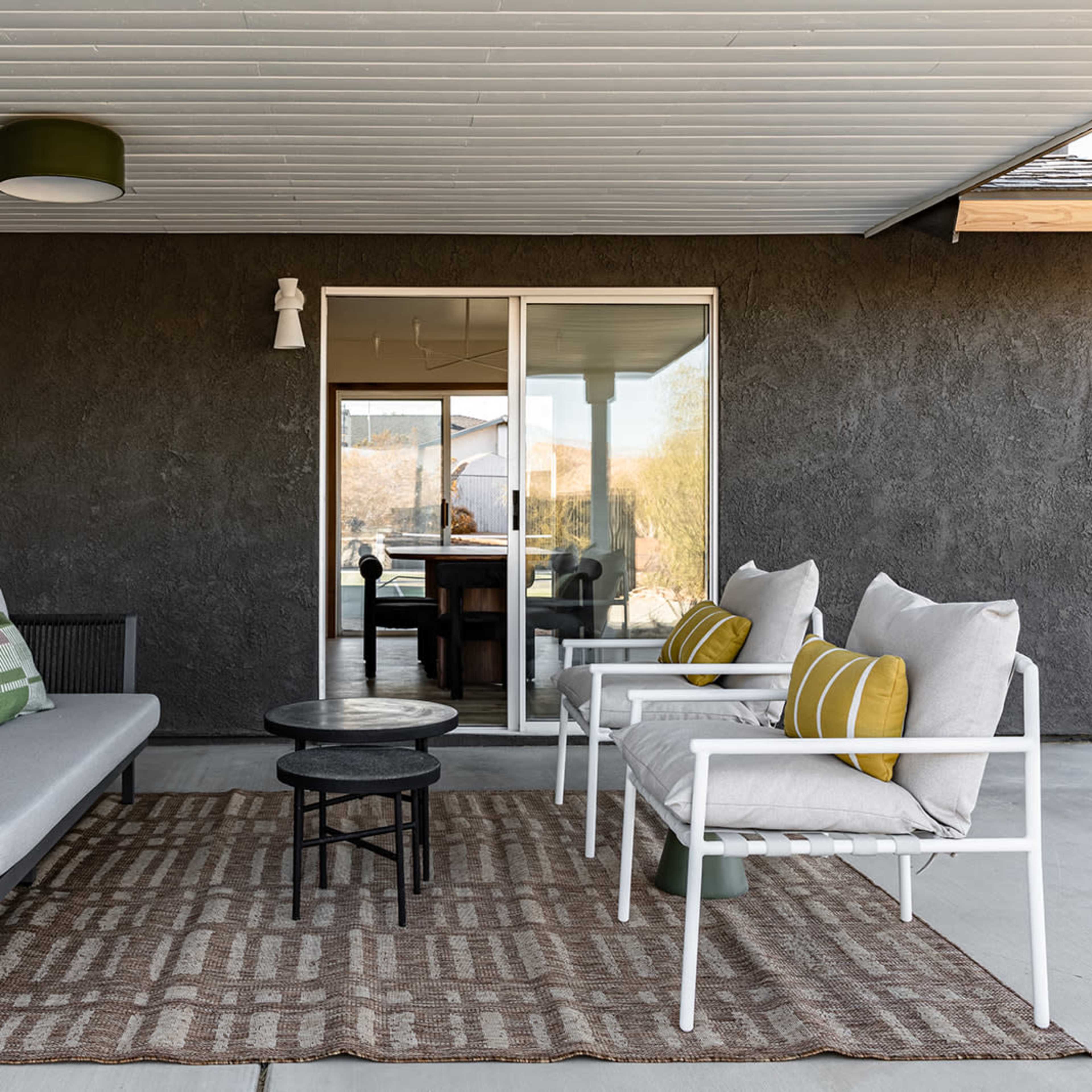 A covered patio features a modern seating arrangement with two chairs and a sofa, set around a small table on a geometric-patterned rug.