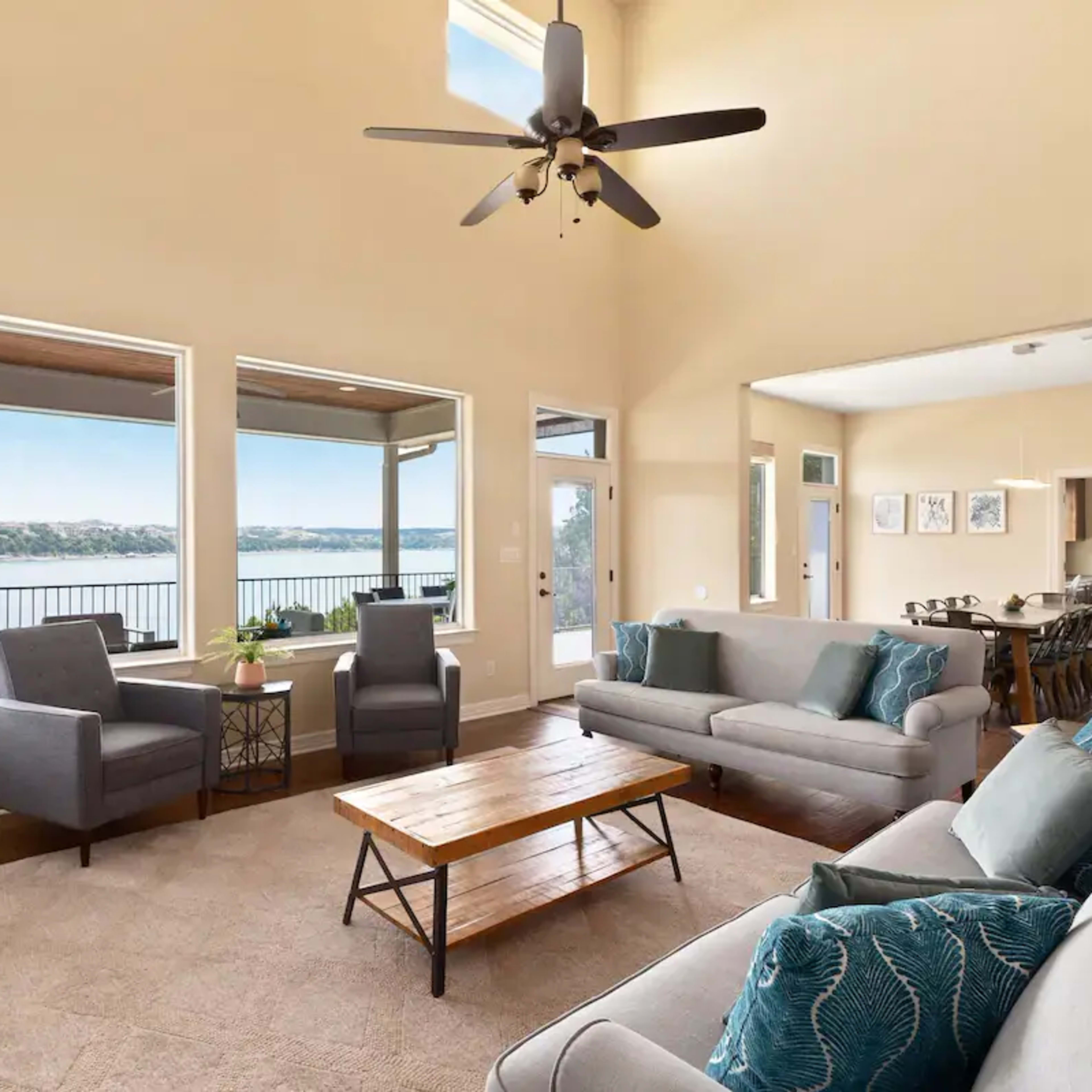 A spacious living room with large windows overlooking a body of water and furnished with a coffee table, two armchairs, and two sofas.