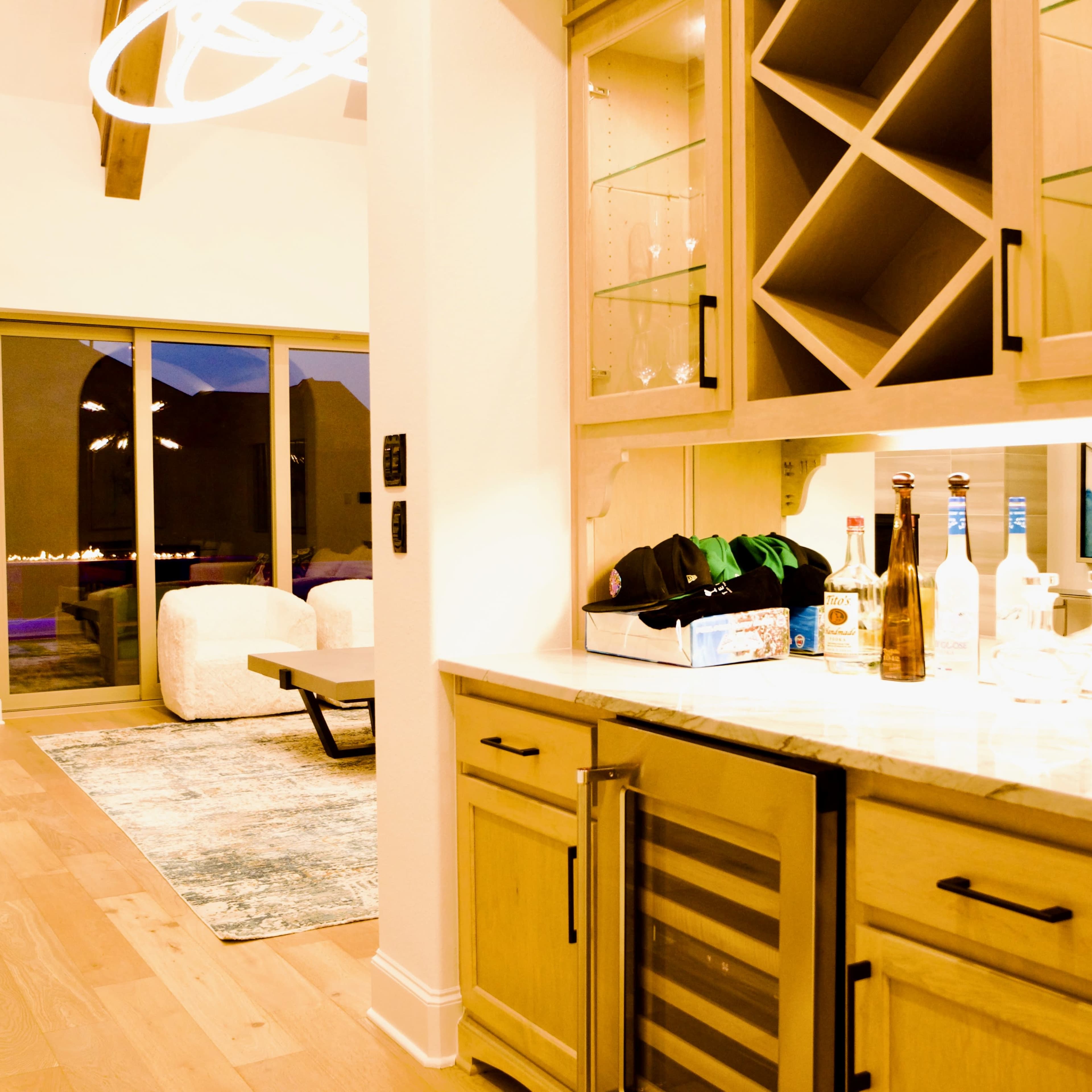 A modern kitchen space with a wine rack, glassware, and several bottles on a countertop, leading to a view of an outdoor area through large glass doors.