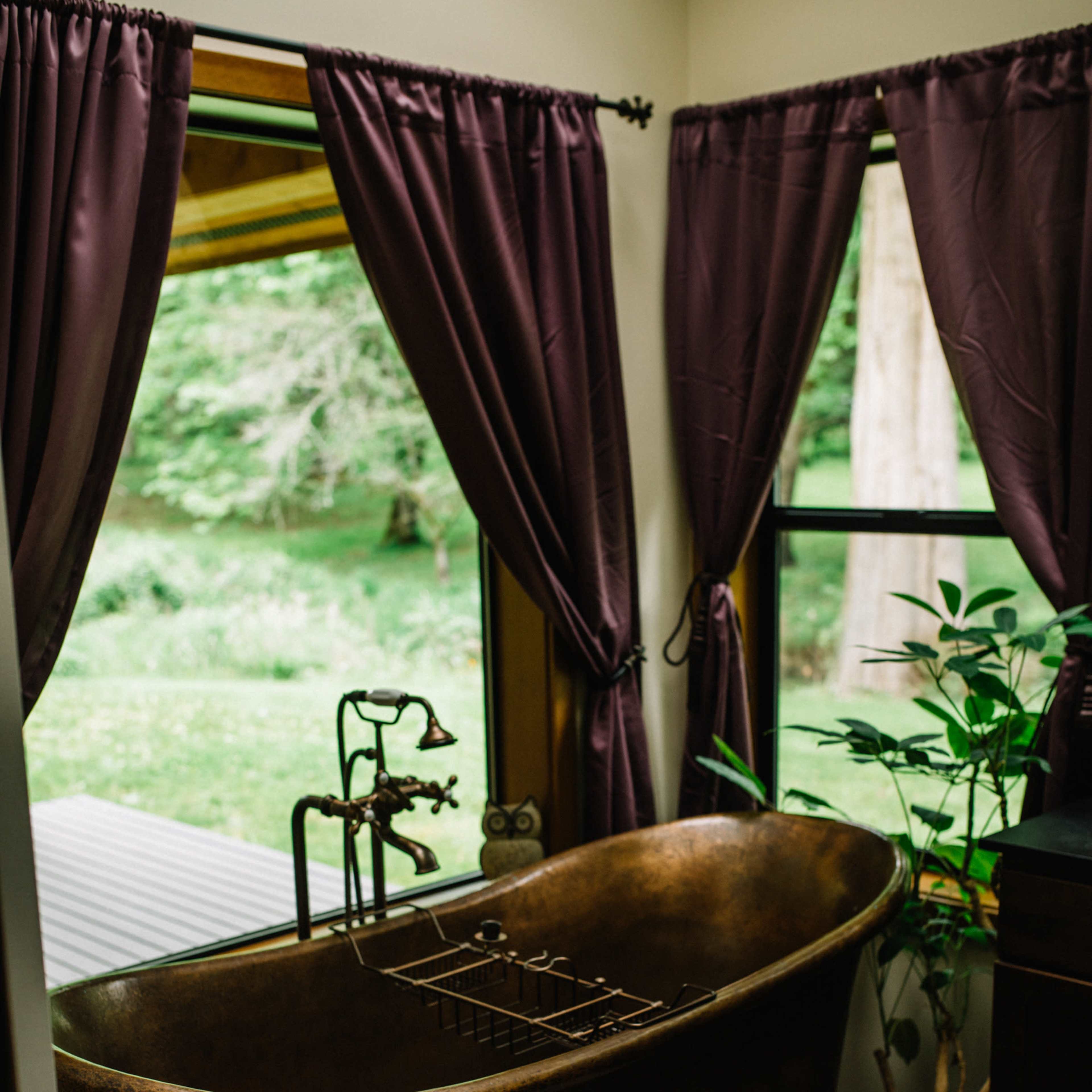 A bronze bathtub with a decorative faucet sits beside a window draped with purple curtains, overlooking a green outdoor landscape.