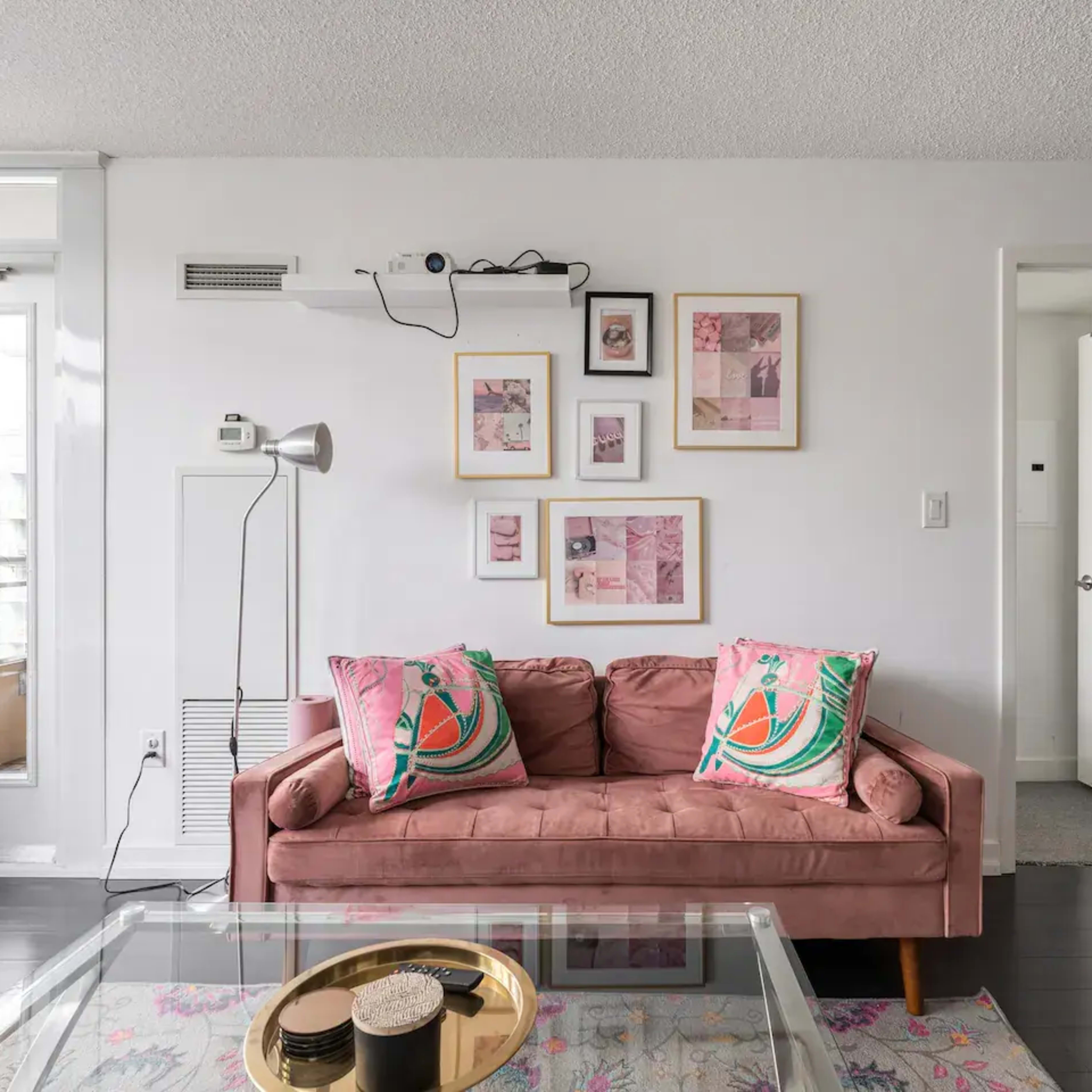 A living room features a pink sofa adorned with patterned cushions, framed artwork on the wall, and a glass coffee table.