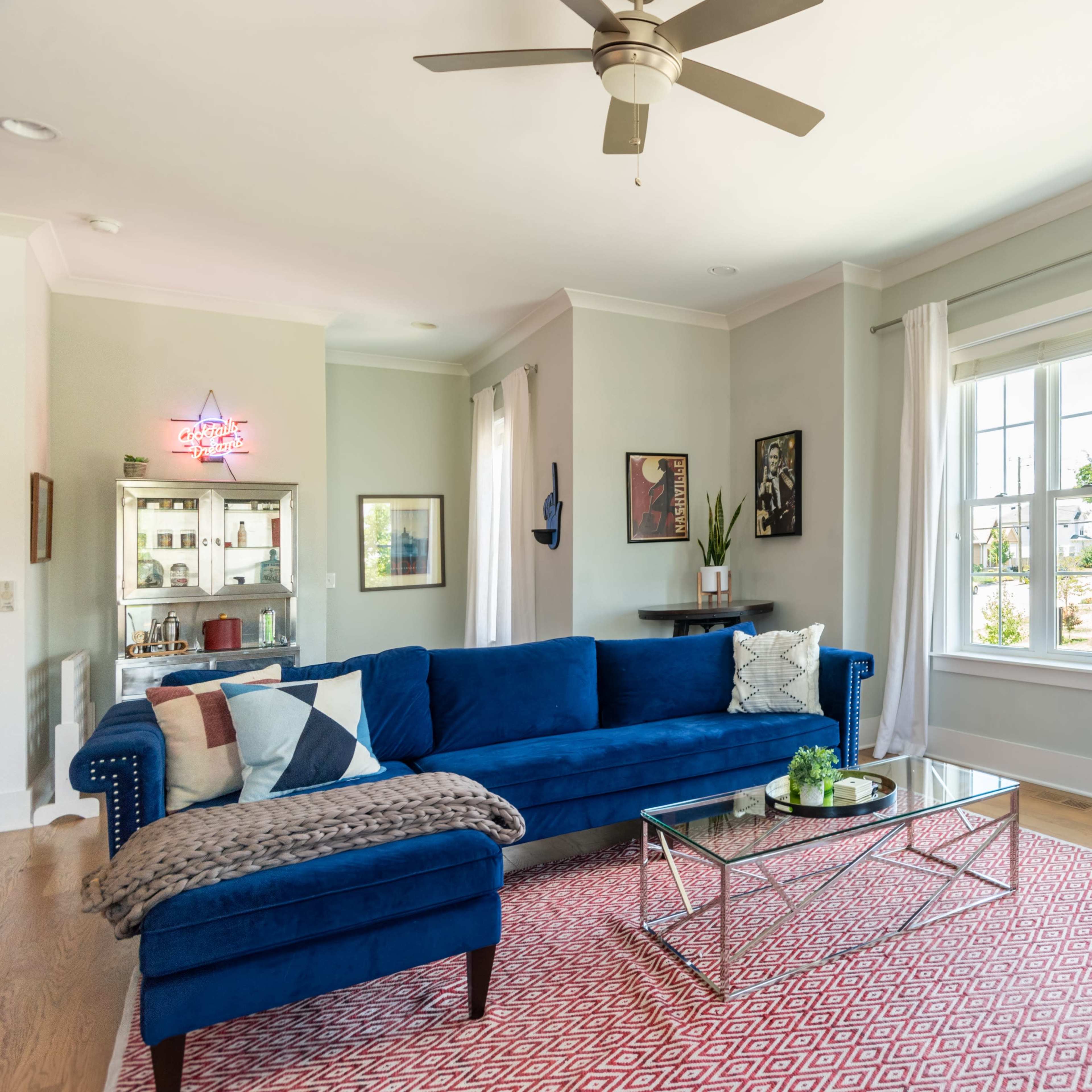 A bright living room features a blue sectional sofa, a glass coffee table, and large windows letting in natural light, with decorative artwork on the walls.