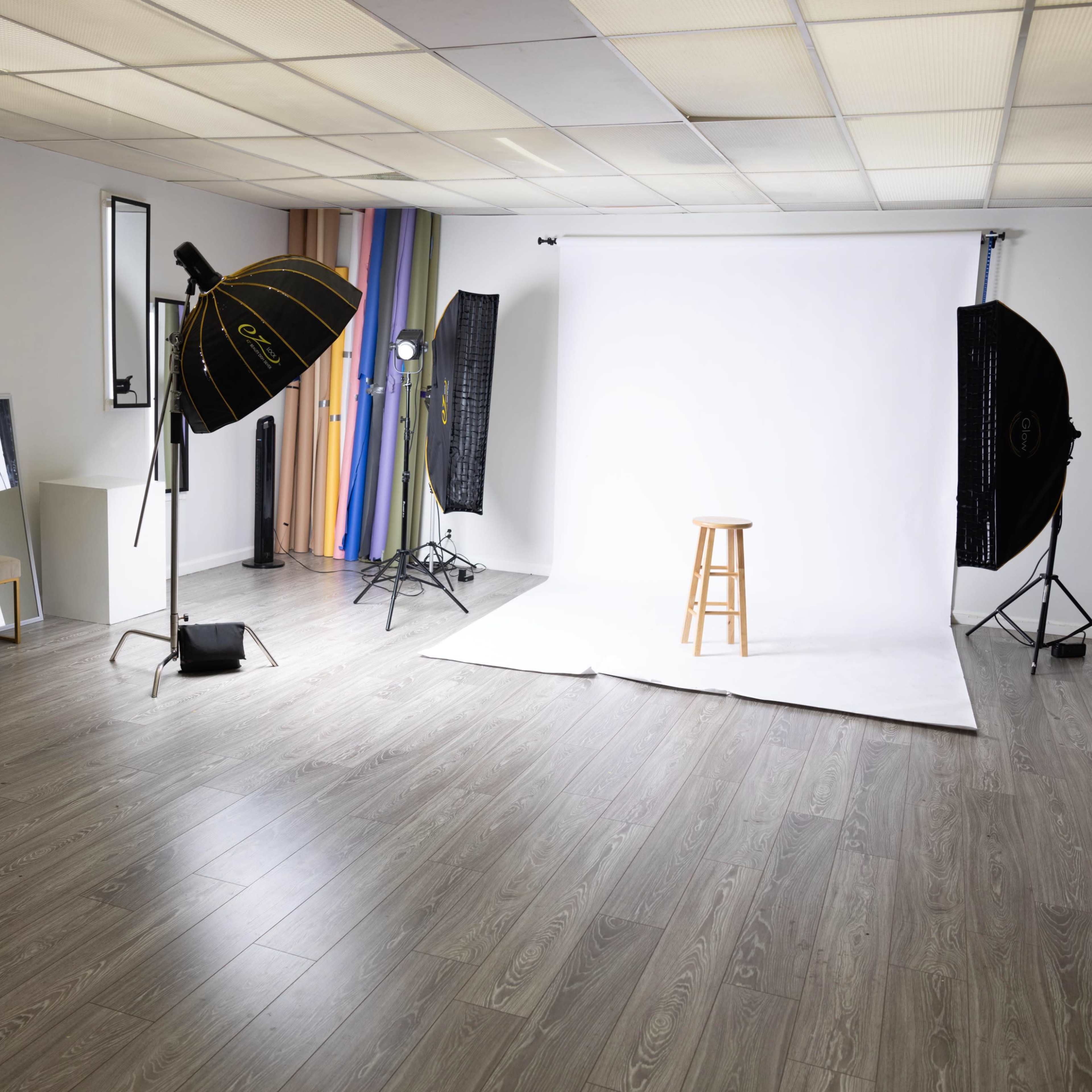 A photography studio with a white backdrop, a wooden stool, and various lighting equipment arranged around the space.