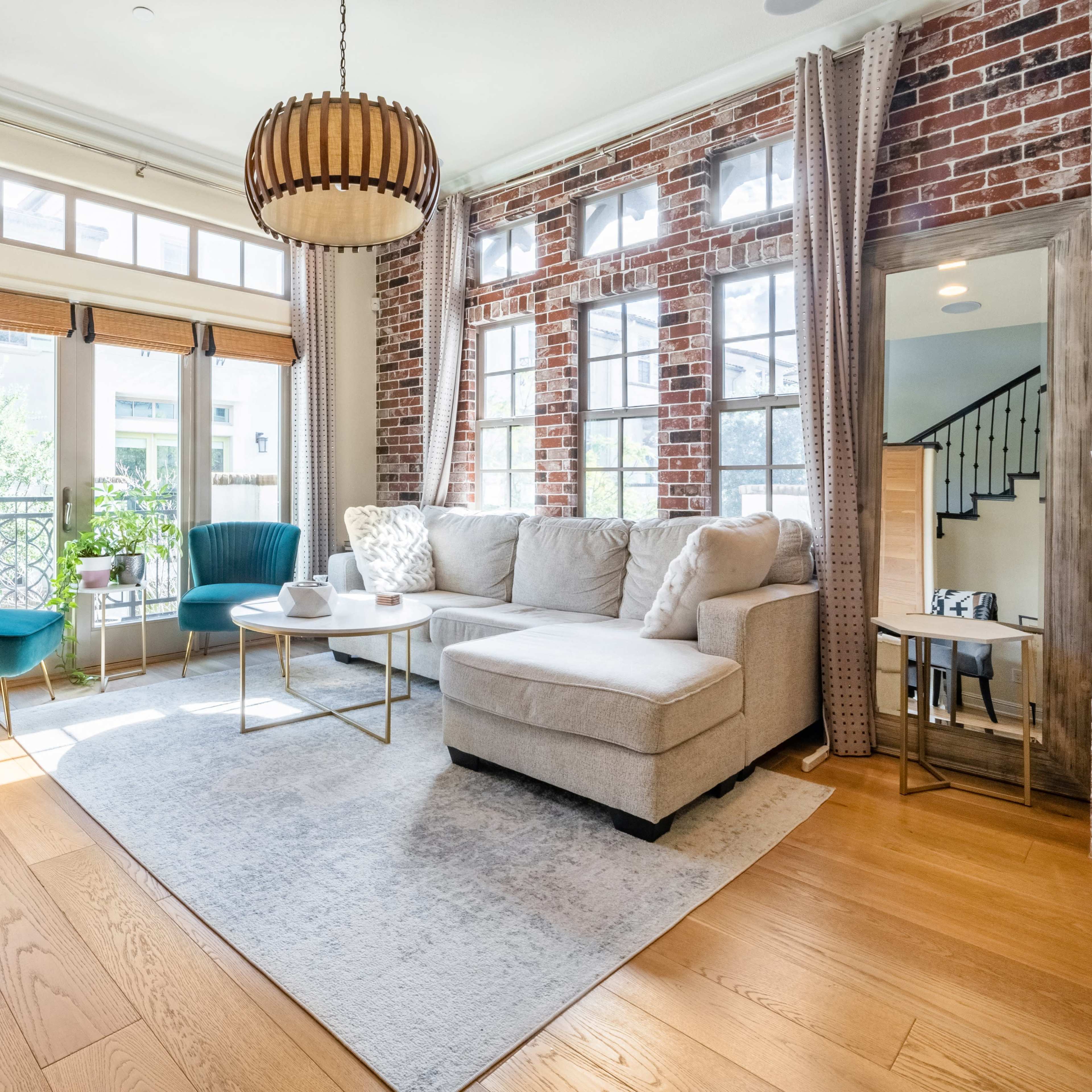 The living room features a beige sectional sofa, a round coffee table, and large windows that provide natural light, with brick walls and green chairs accentuating the decor.