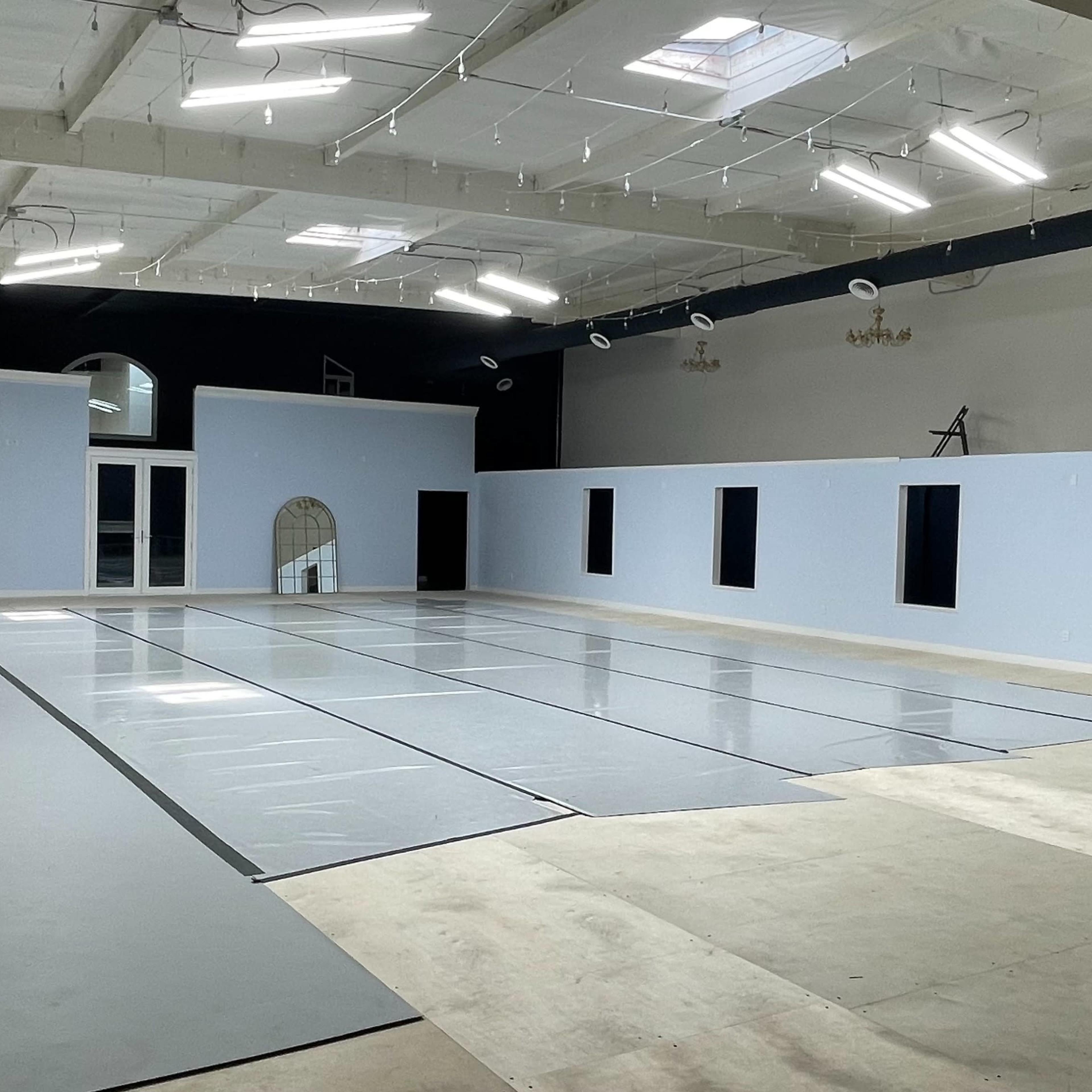 A spacious, well-lit room with empty gray flooring and light blue walls, featuring several windows and a large open area.