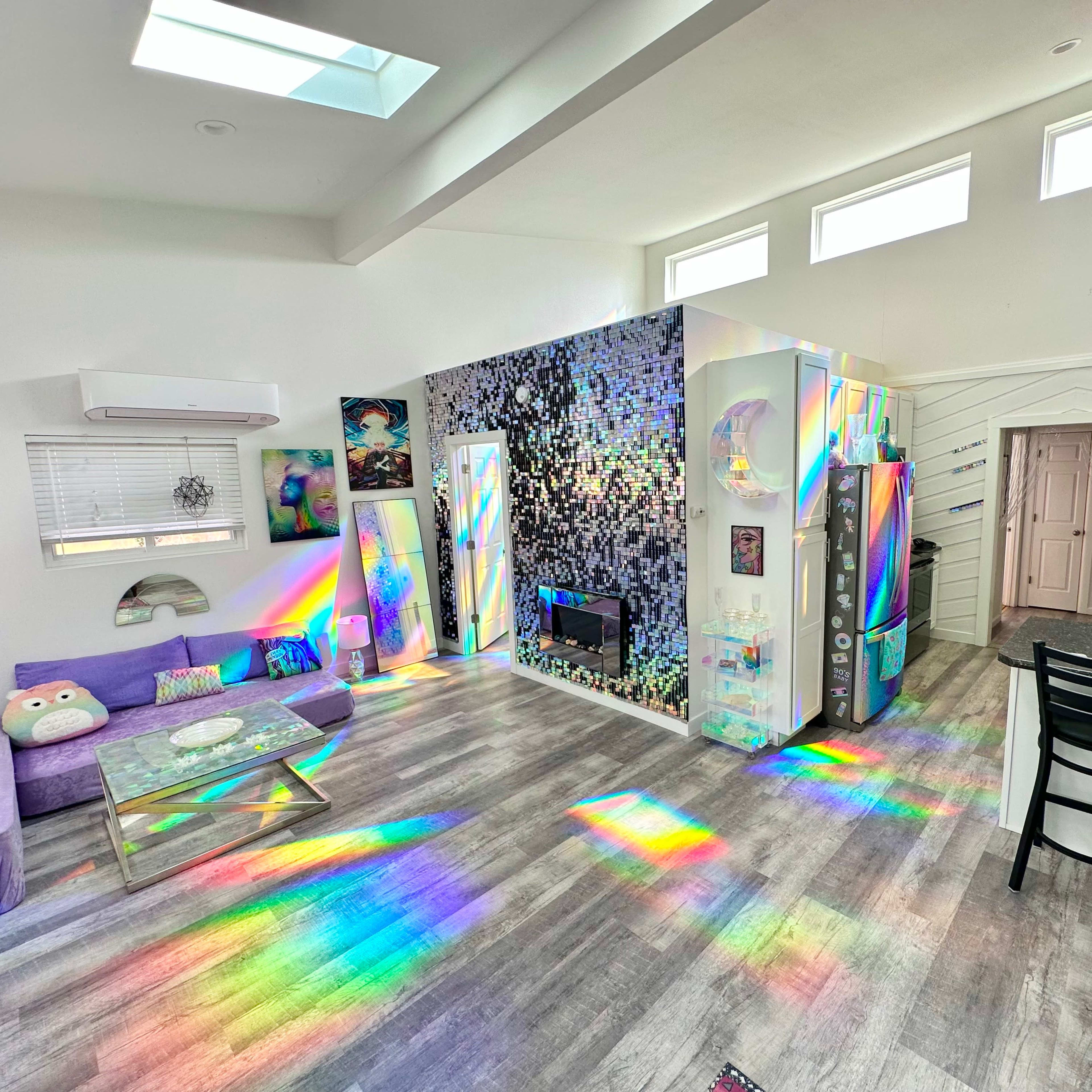 A brightly lit living space featuring colorful artwork, a modern kitchen, and walls adorned with reflective surfaces creating rainbow light patterns on the floor.