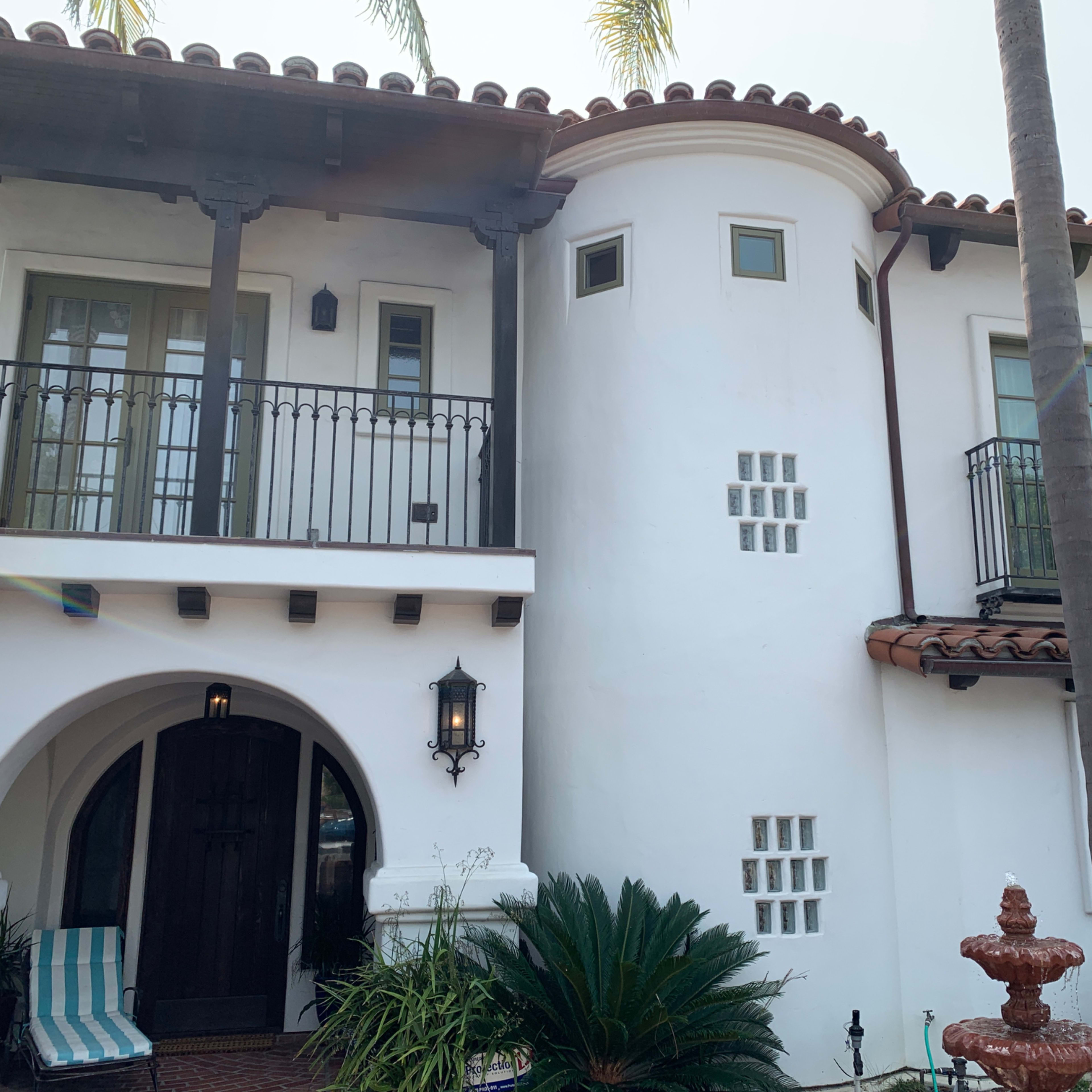 A two-story white stucco house with a rounded corner and balconies, flanked by palm trees and featuring a front entrance with a wooden door.