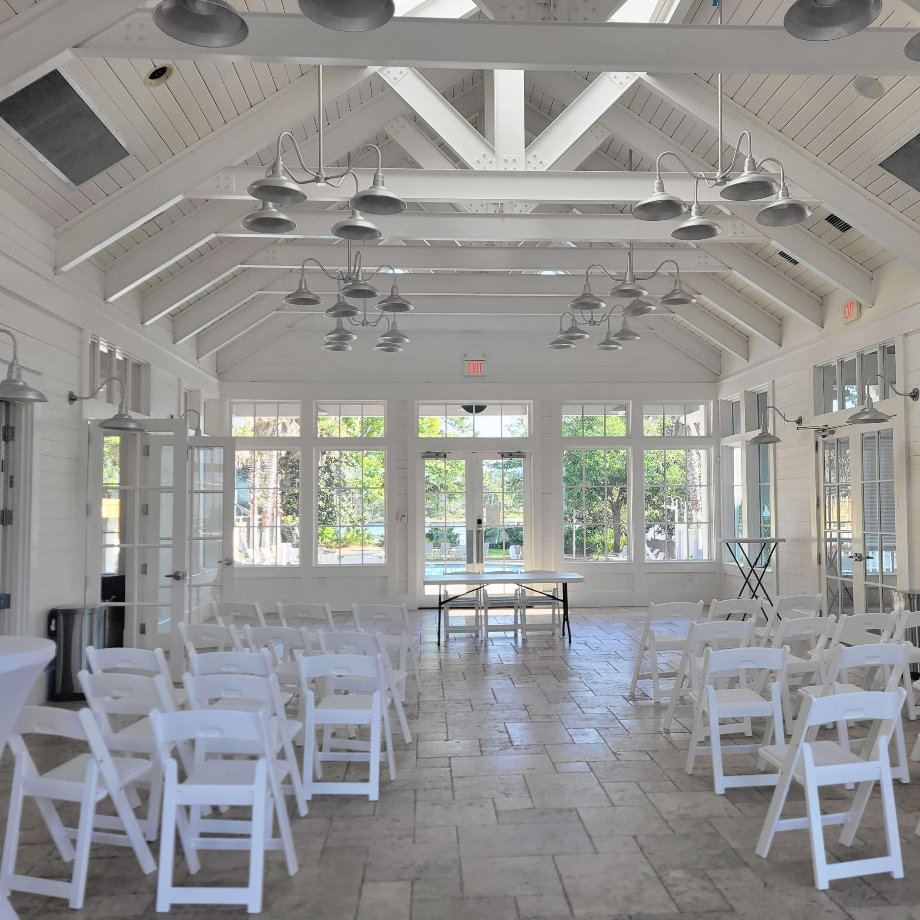 A bright, spacious room with white walls and a vaulted ceiling, set up with rows of white chairs facing a table at the front.