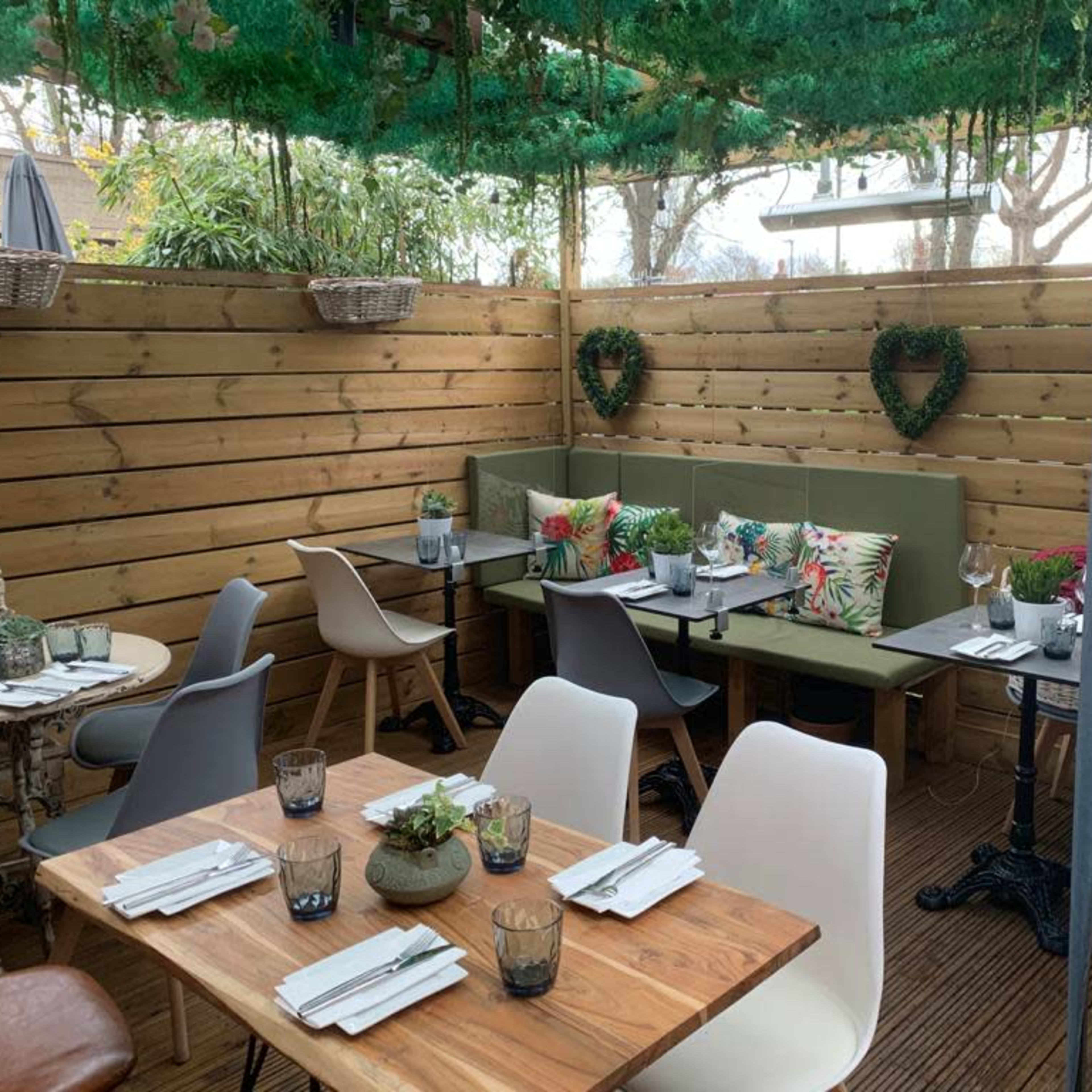 A cozy outdoor dining area with wooden walls, various tables, and seating arrangements adorned with plants and cushions.