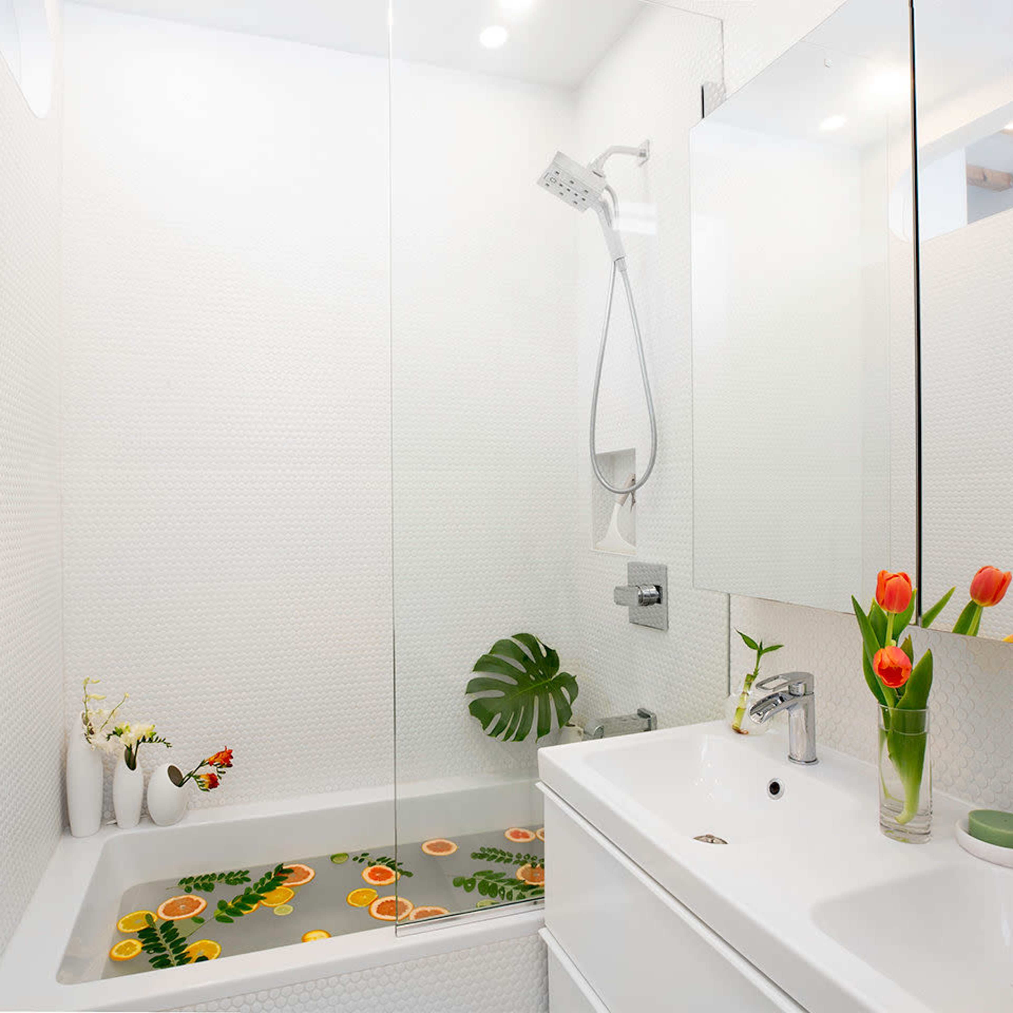 A modern bathroom with a bathtub filled with water and orange slices, surrounded by white tiles and a small flower arrangement.