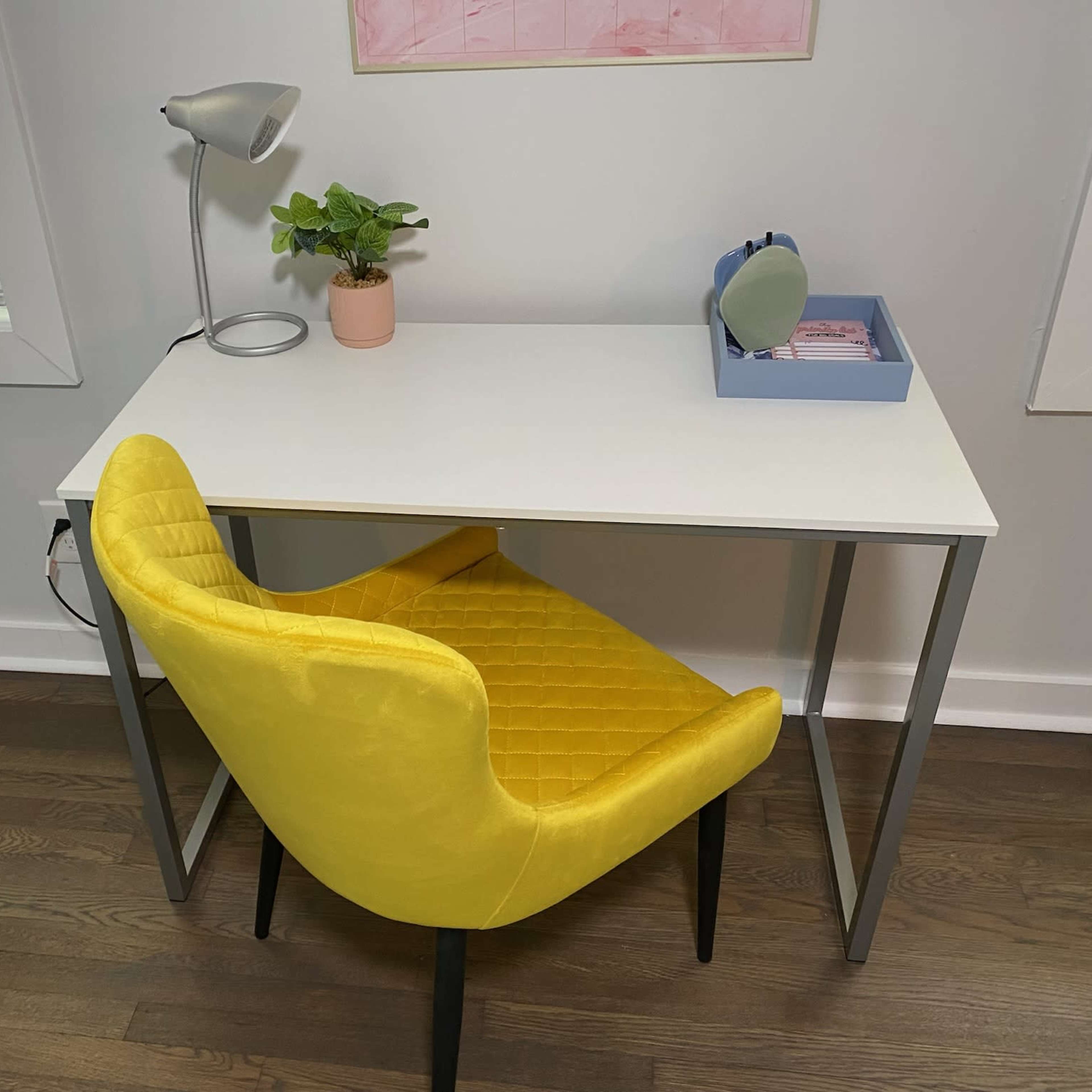 A white desk with a yellow upholstered chair, a small potted plant, and a lamp is positioned against a wall with partially open blinds.