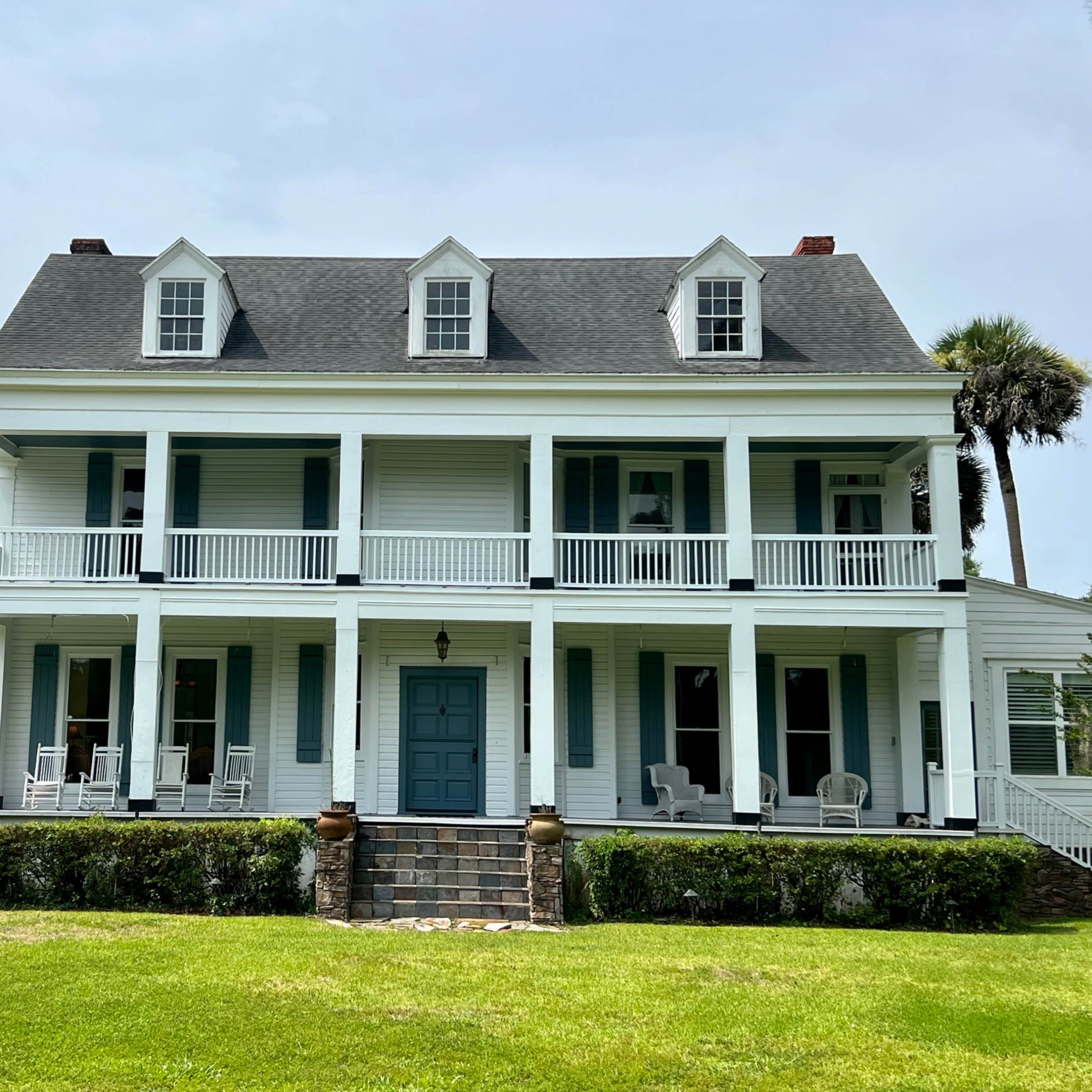 A large, two-story white house with a blue front door and a porch supported by columns is set on a grassy lawn.