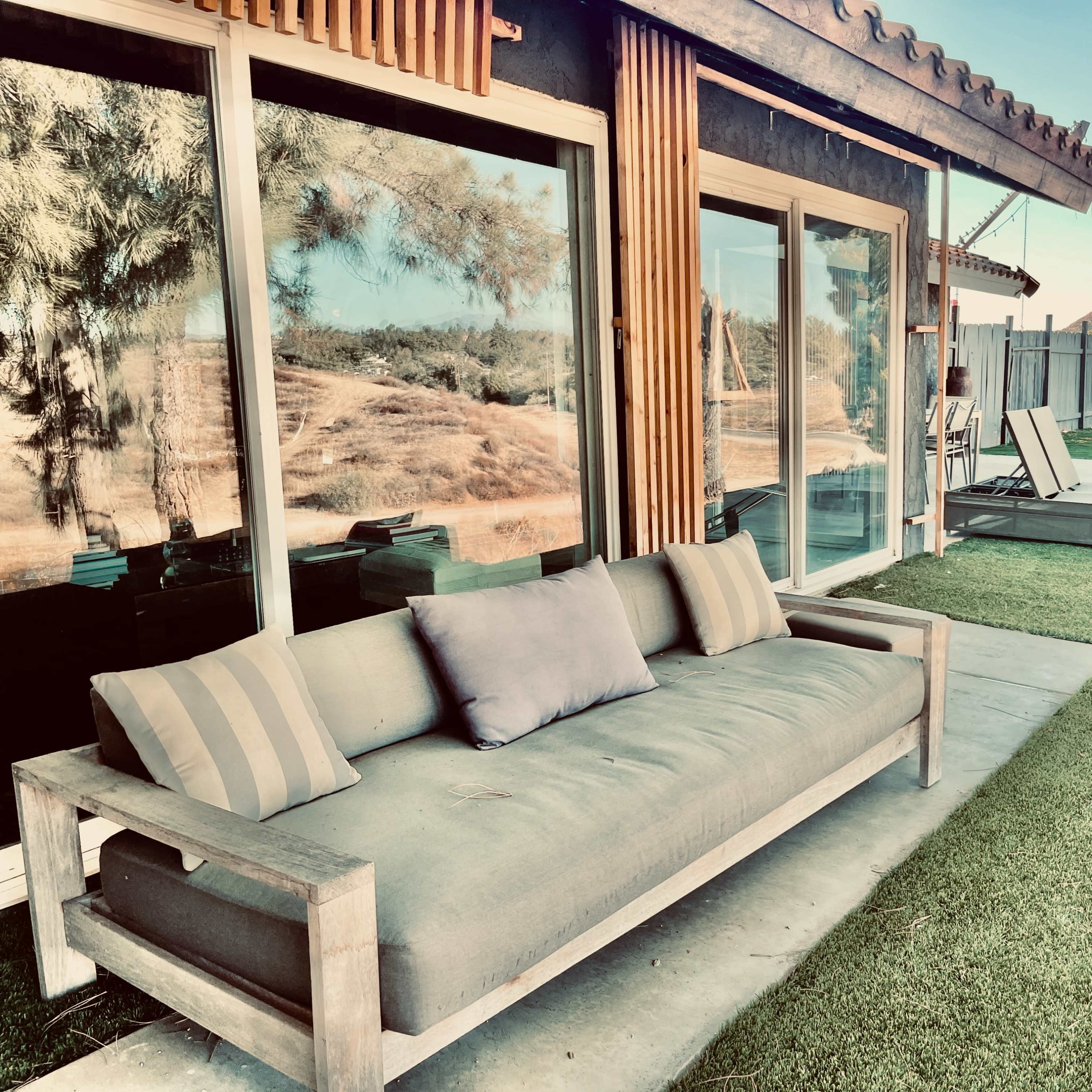 A modern outdoor patio features a wooden sofa with cushions and lounge chairs, overlooking a grassy area and distant hills.