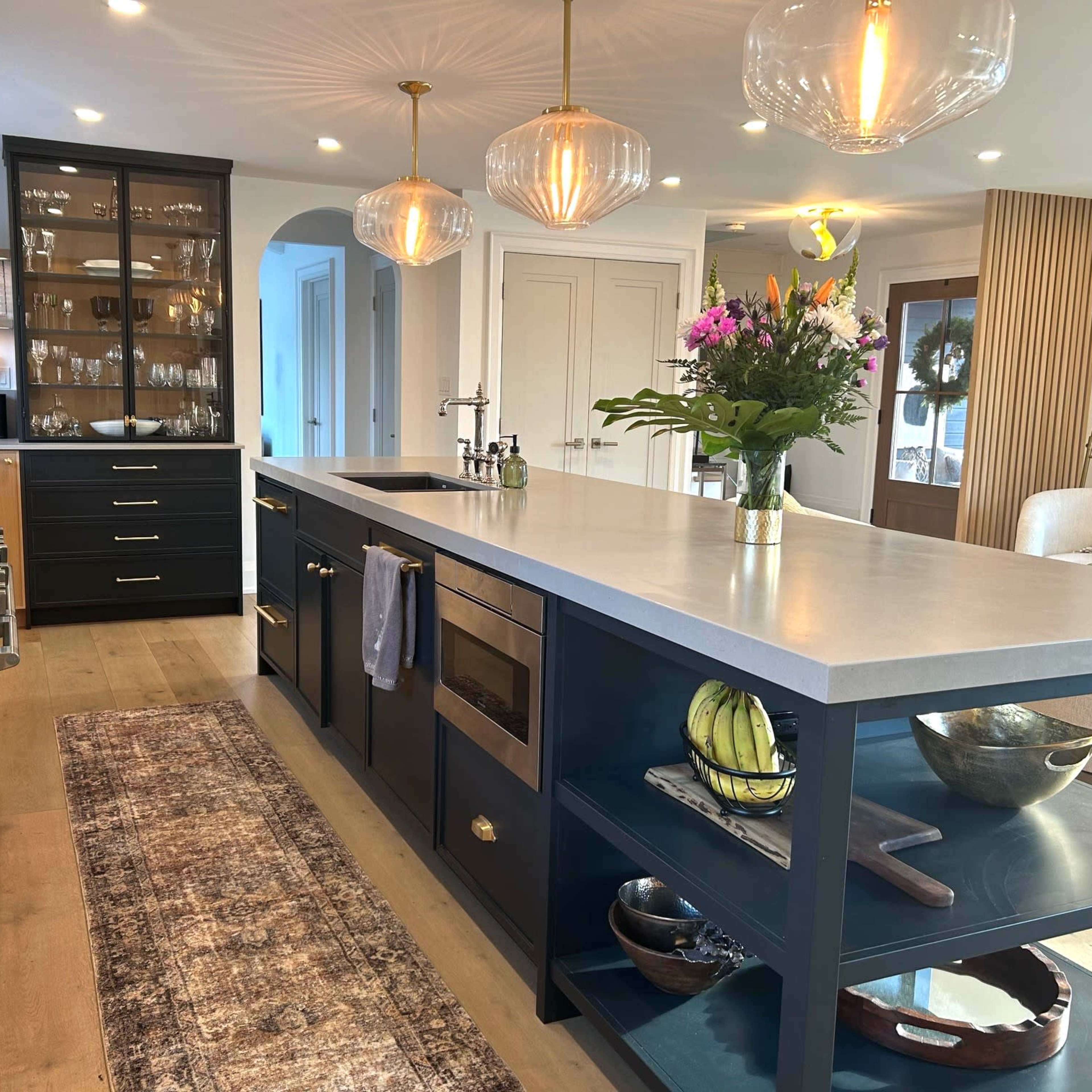 A modern kitchen features a central island with a countertop, a gas stove, dark cabinetry, glass-front shelves displaying dishware, and decorative plants.