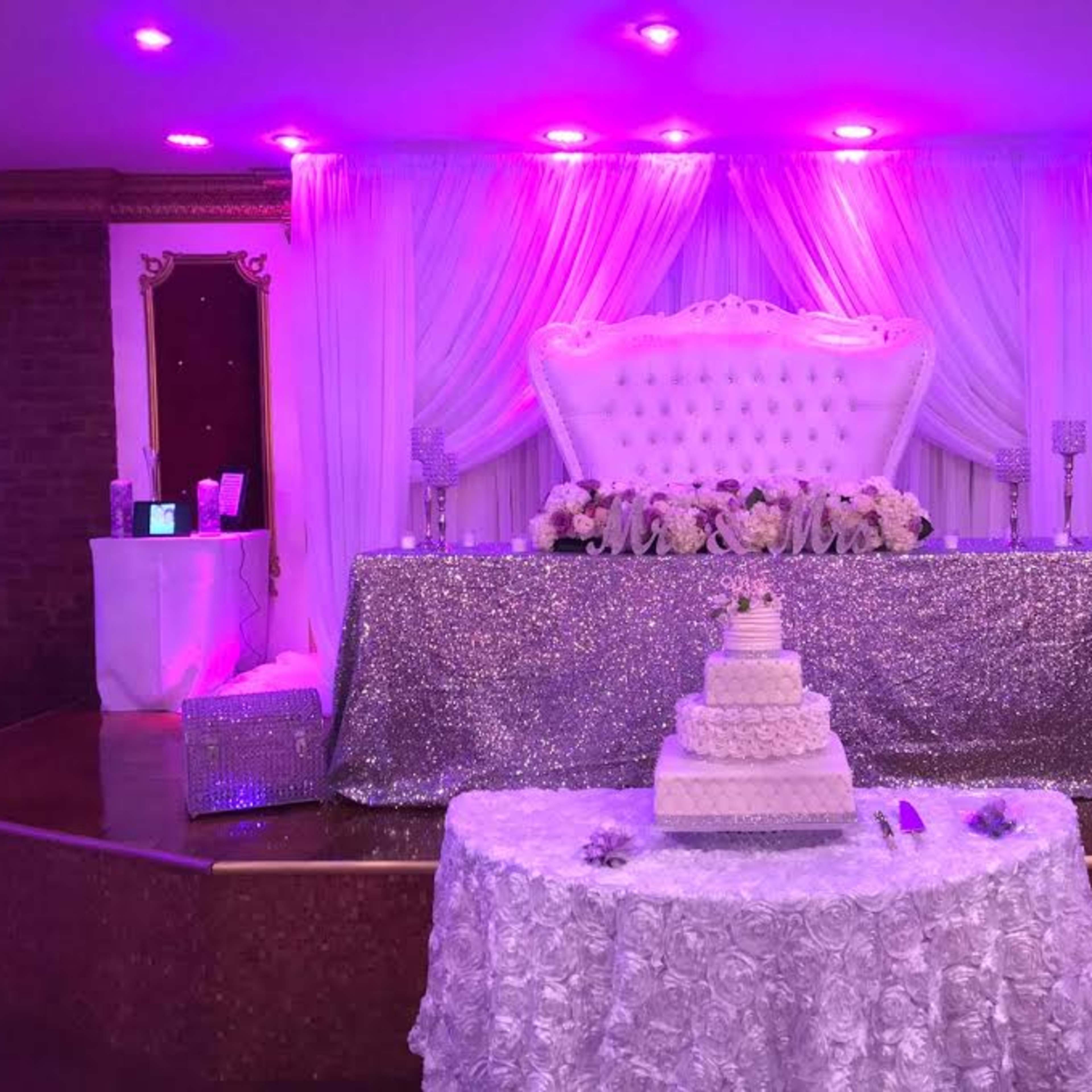 A decorated event space with a white and silver cake on a lace tablecloth in front of a lavish backdrop featuring a large tufted white settee and draped fabric illuminated by purple lighting.