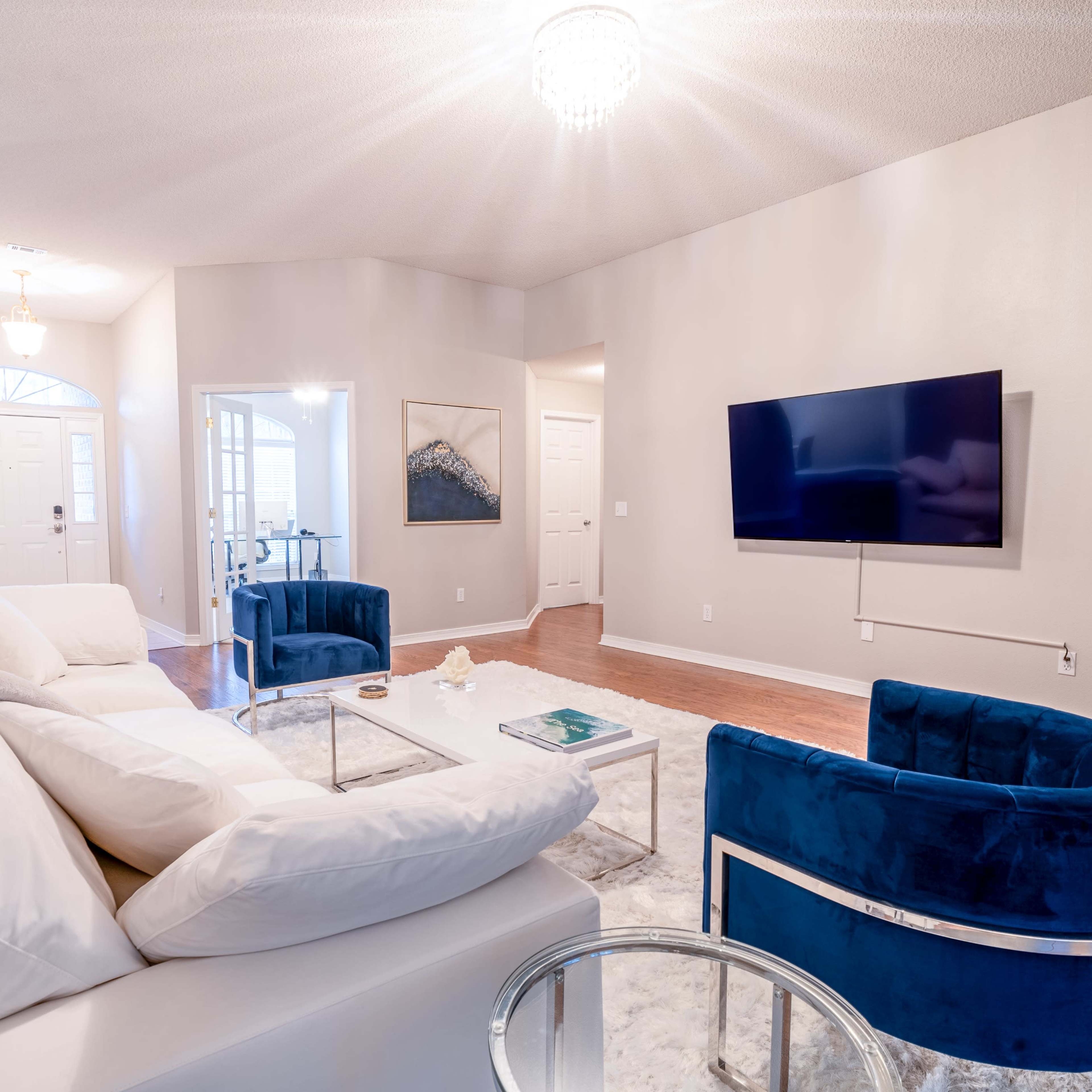 A modern living room features a white sectional sofa, two blue velvet chairs, a glass coffee table, and a wall-mounted television.