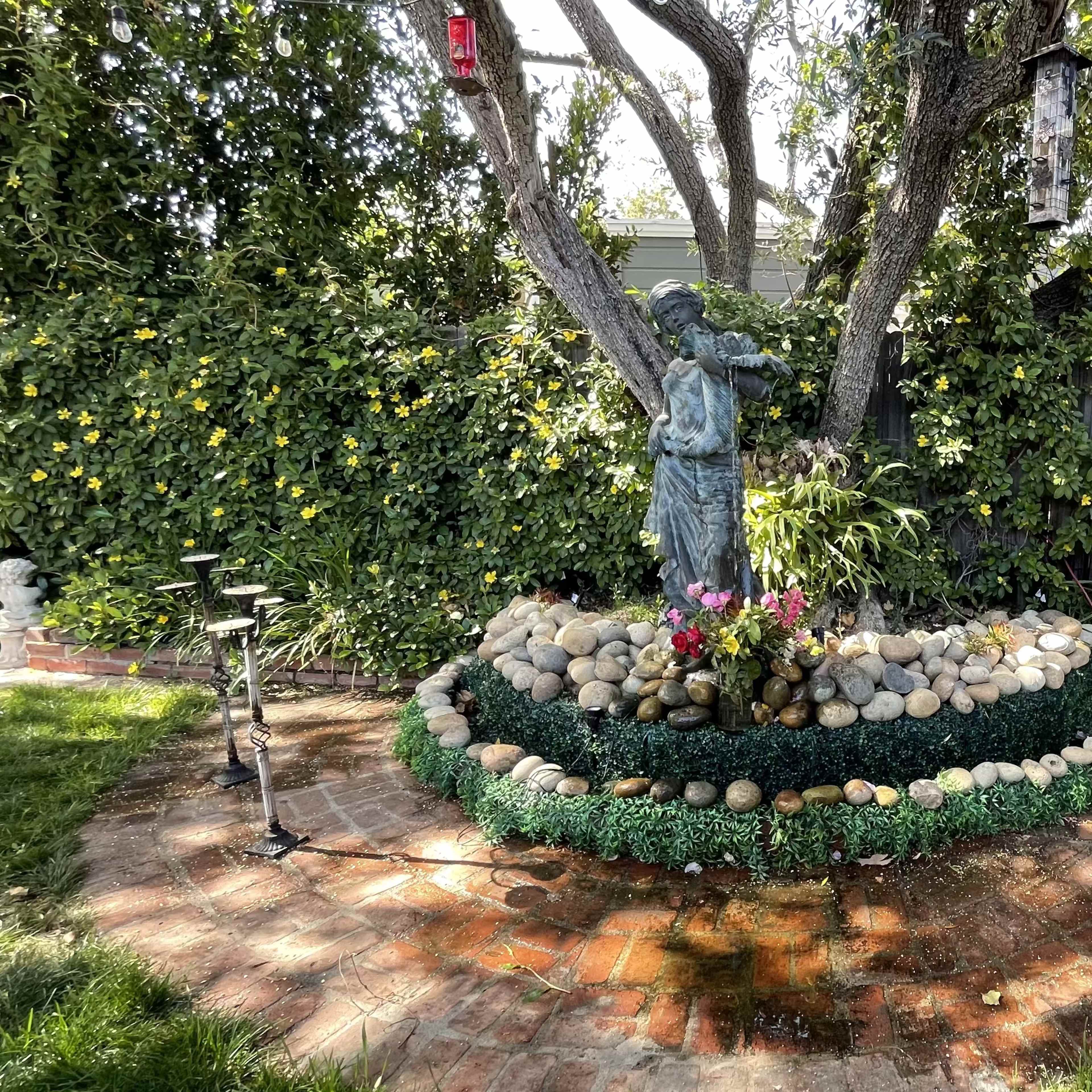 A landscaped garden featuring a statue surrounded by rocks and flowering plants, situated under trees.