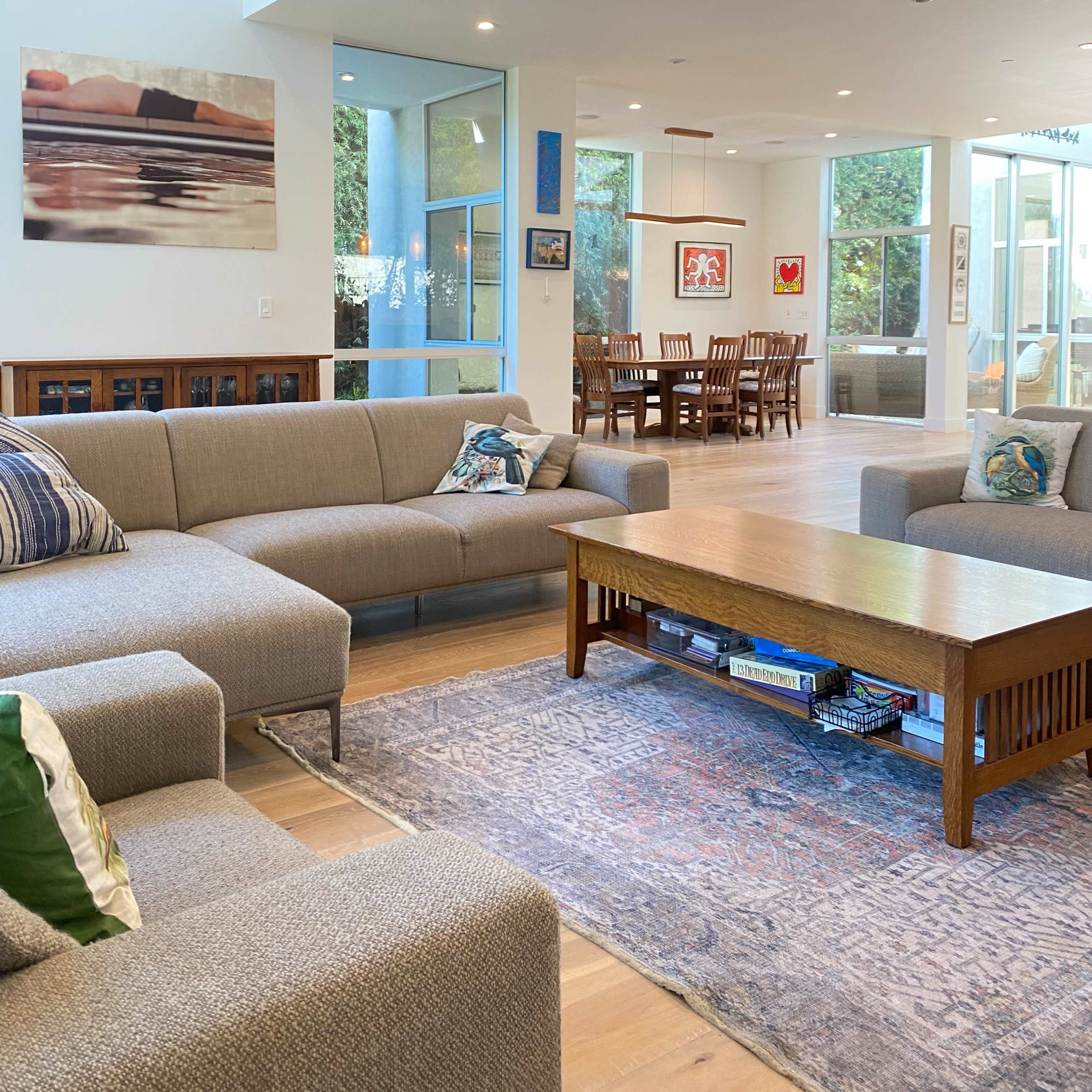 A modern living room with a light gray sectional sofa, a wooden coffee table, and a dining area in the background that includes a wooden dining table and chairs.