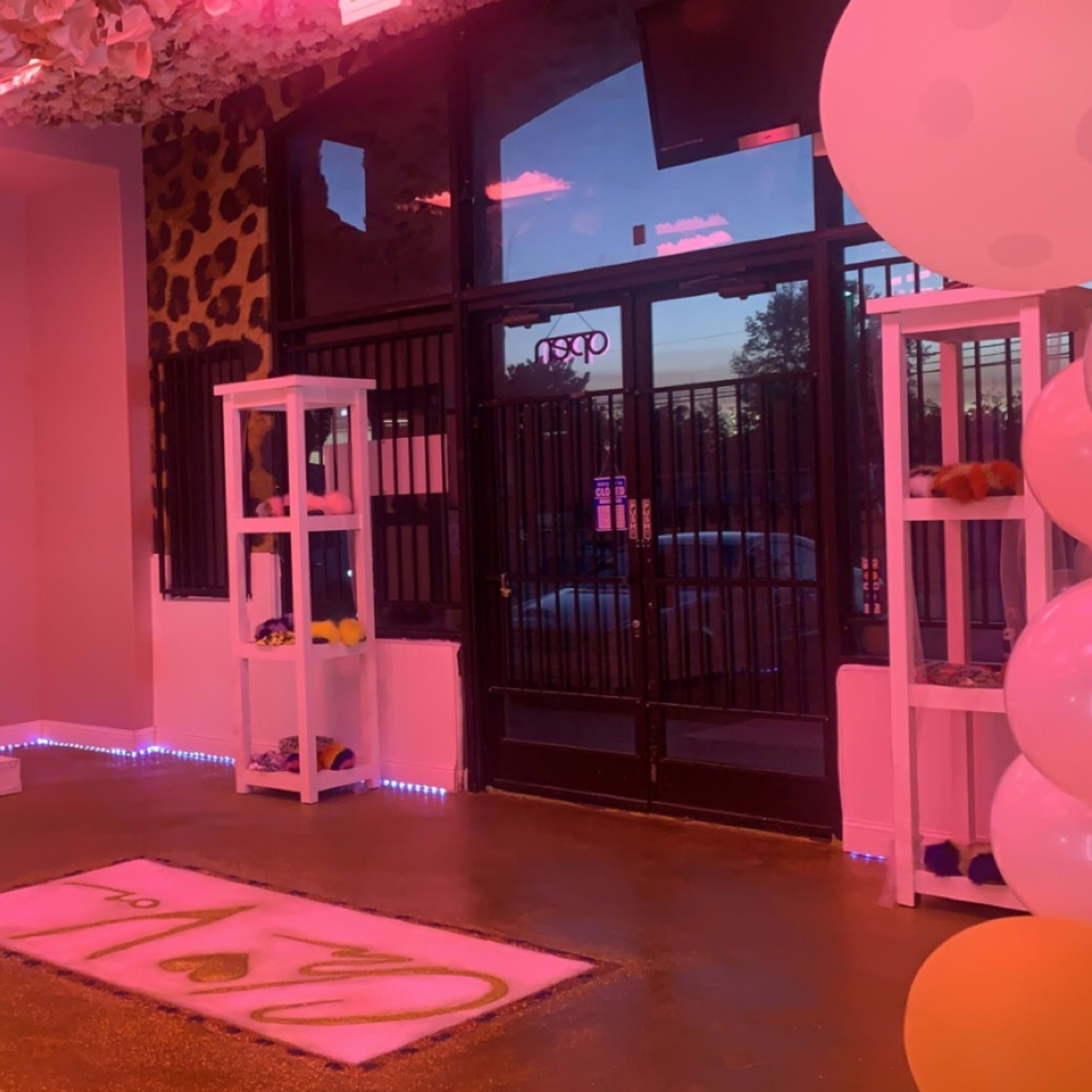 An indoor space with pink lighting, featuring a reception area, shelves displaying items, and balloons near the entrance.