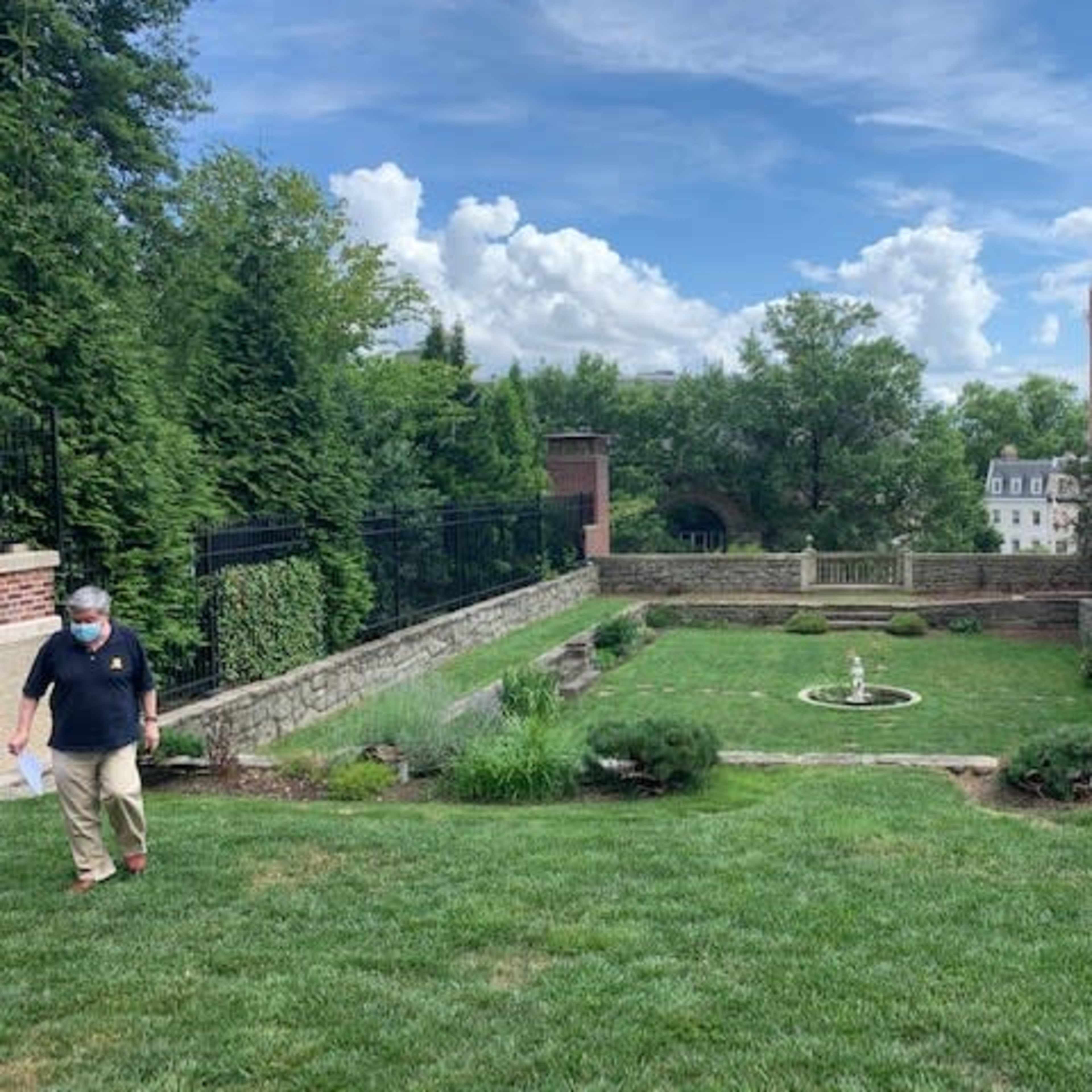 A man walks through a green lawn with a fountain and landscaped garden surrounded by trees and buildings under a partly cloudy sky.