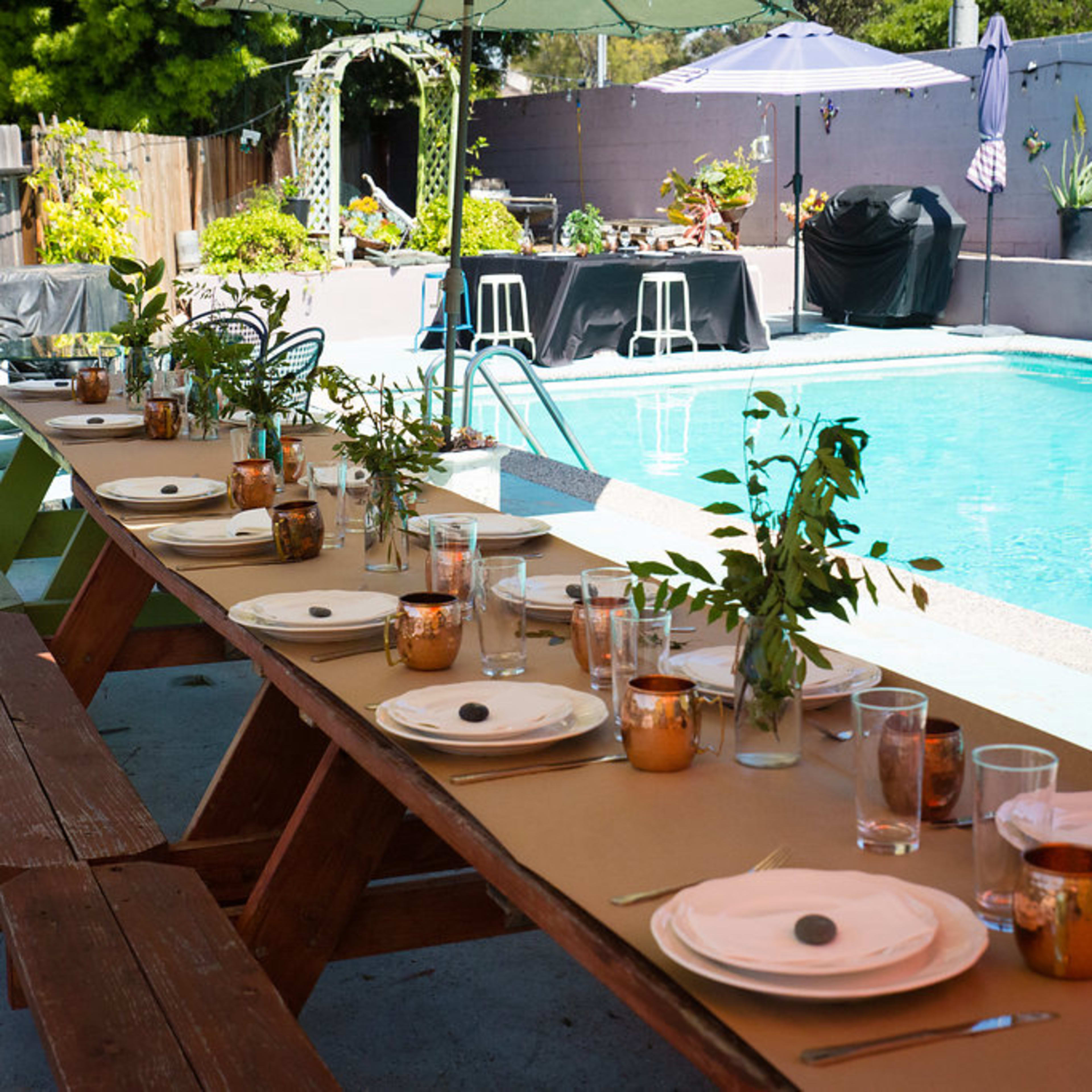 A long wooden table is set with dishes and glasses beside a swimming pool in a backyard garden.