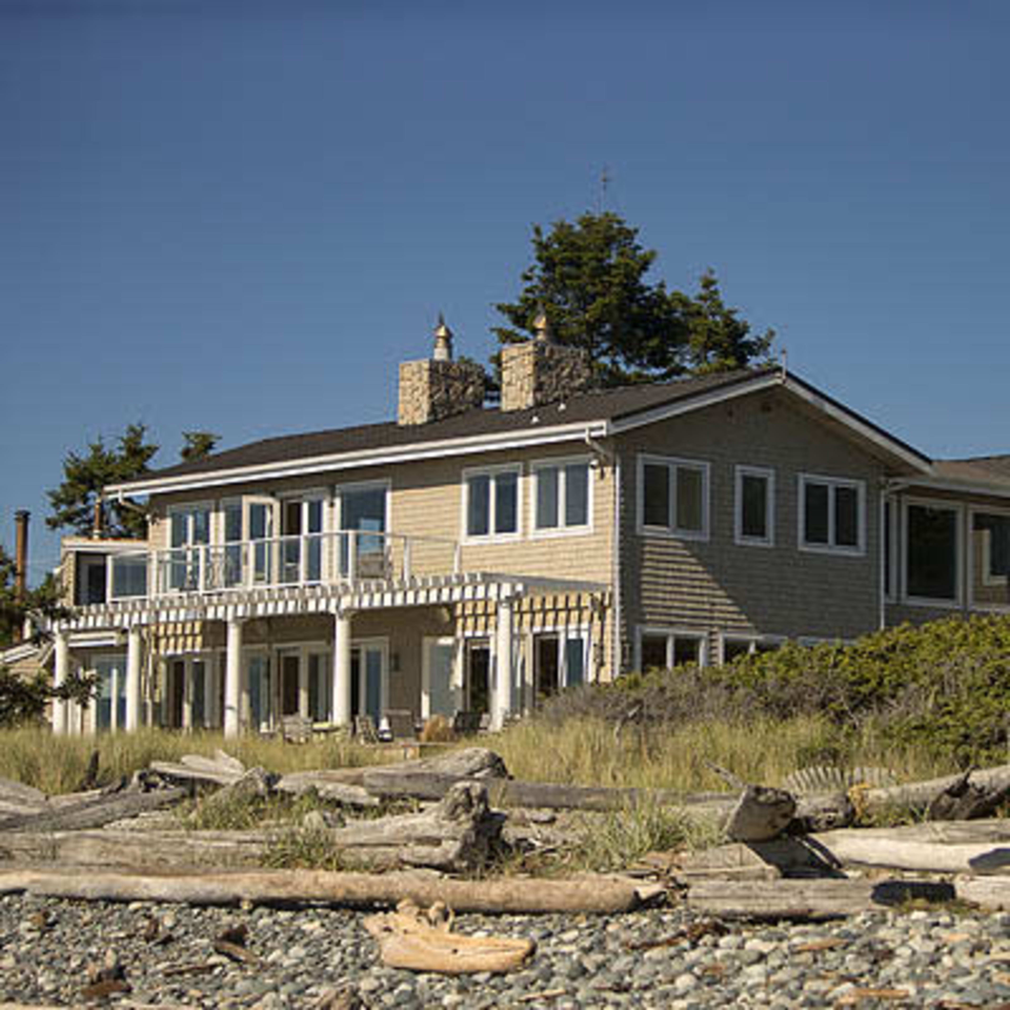 A large beach house with multiple levels and a wooden deck sits near the shore, surrounded by sand and driftwood.