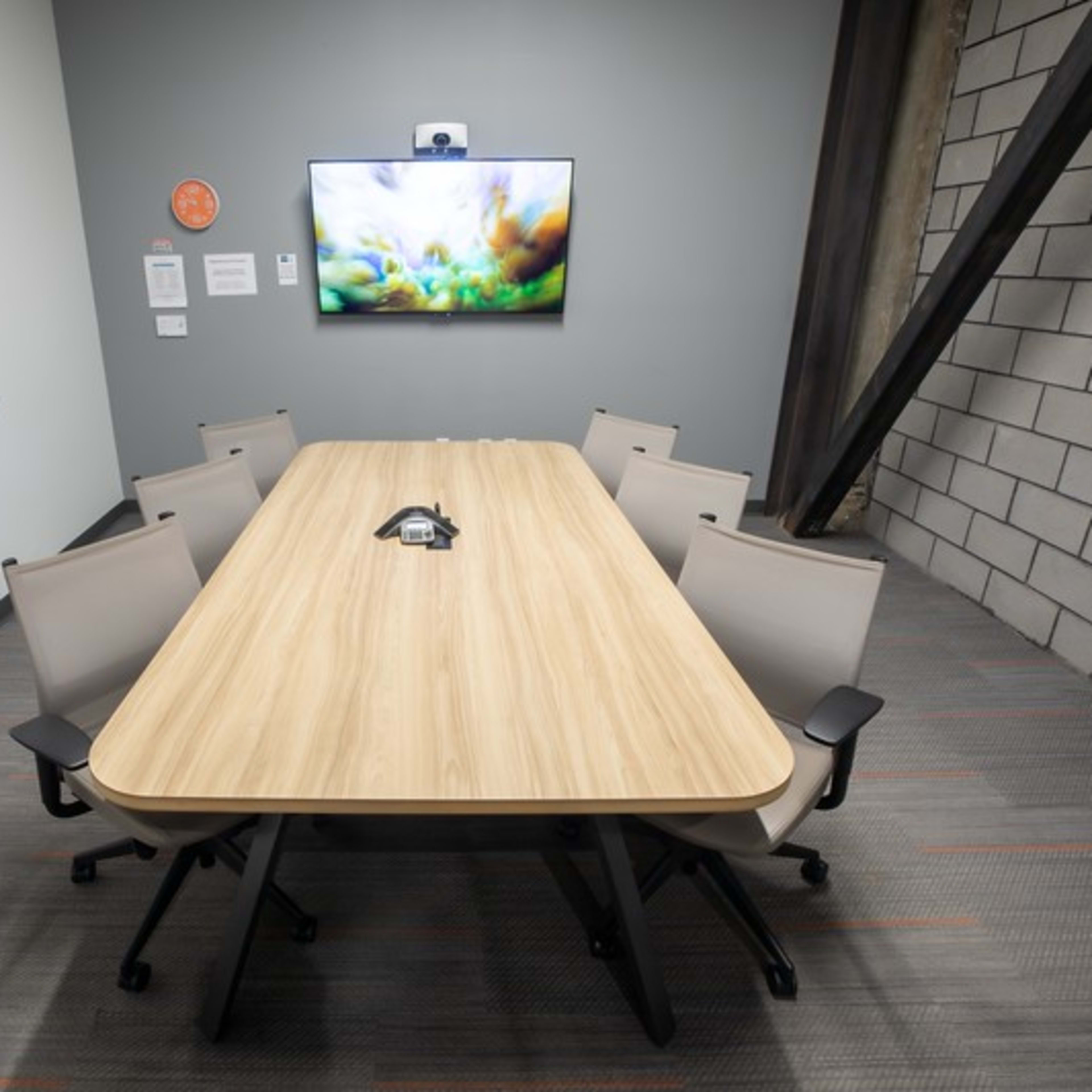 A conference room features a long wooden table surrounded by six chairs, with a wall-mounted screen displaying a colorful image.