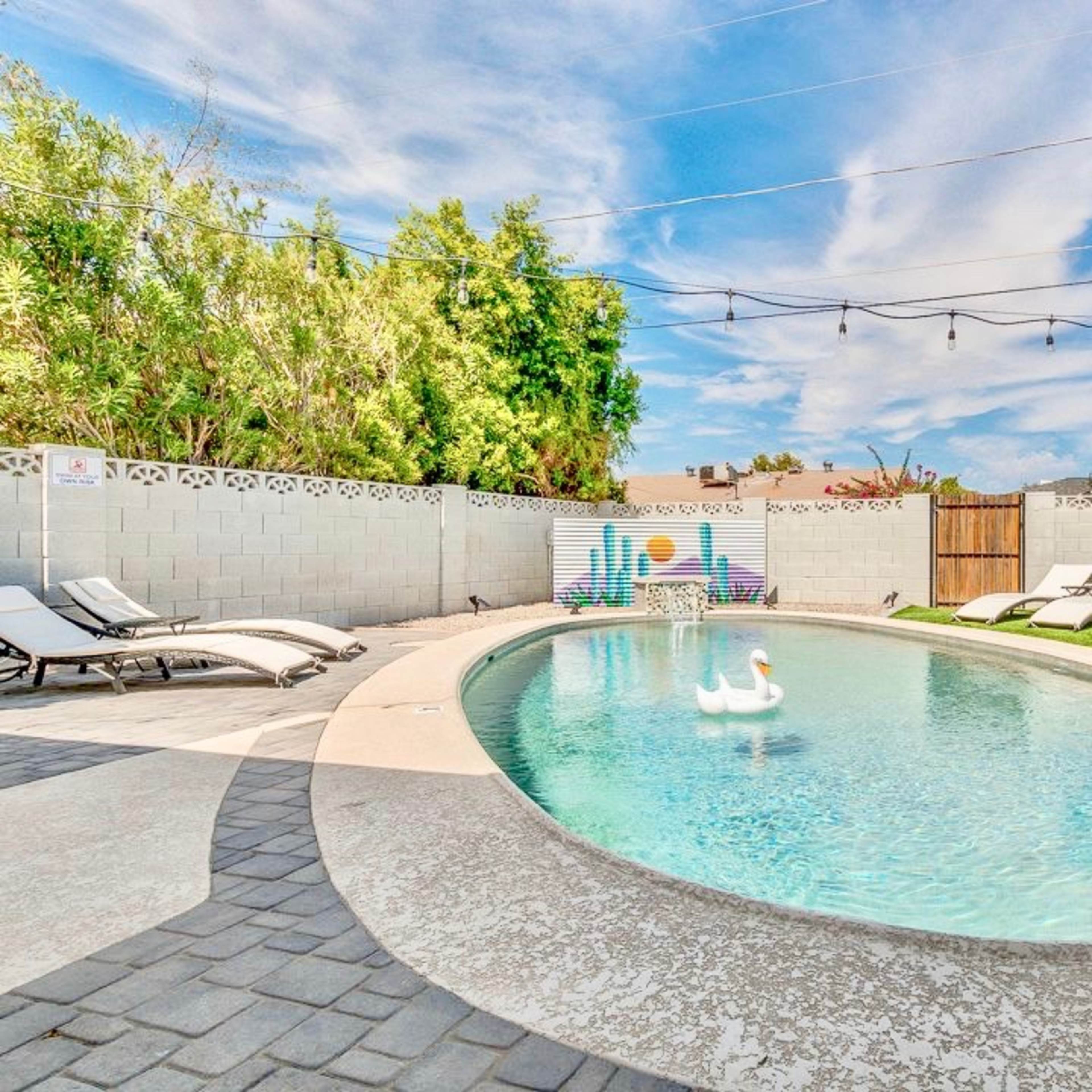 A backyard pool area features a clear water pool with a large inflatable swan and rubber ducks, surrounded by lounge chairs and decorative string lights.